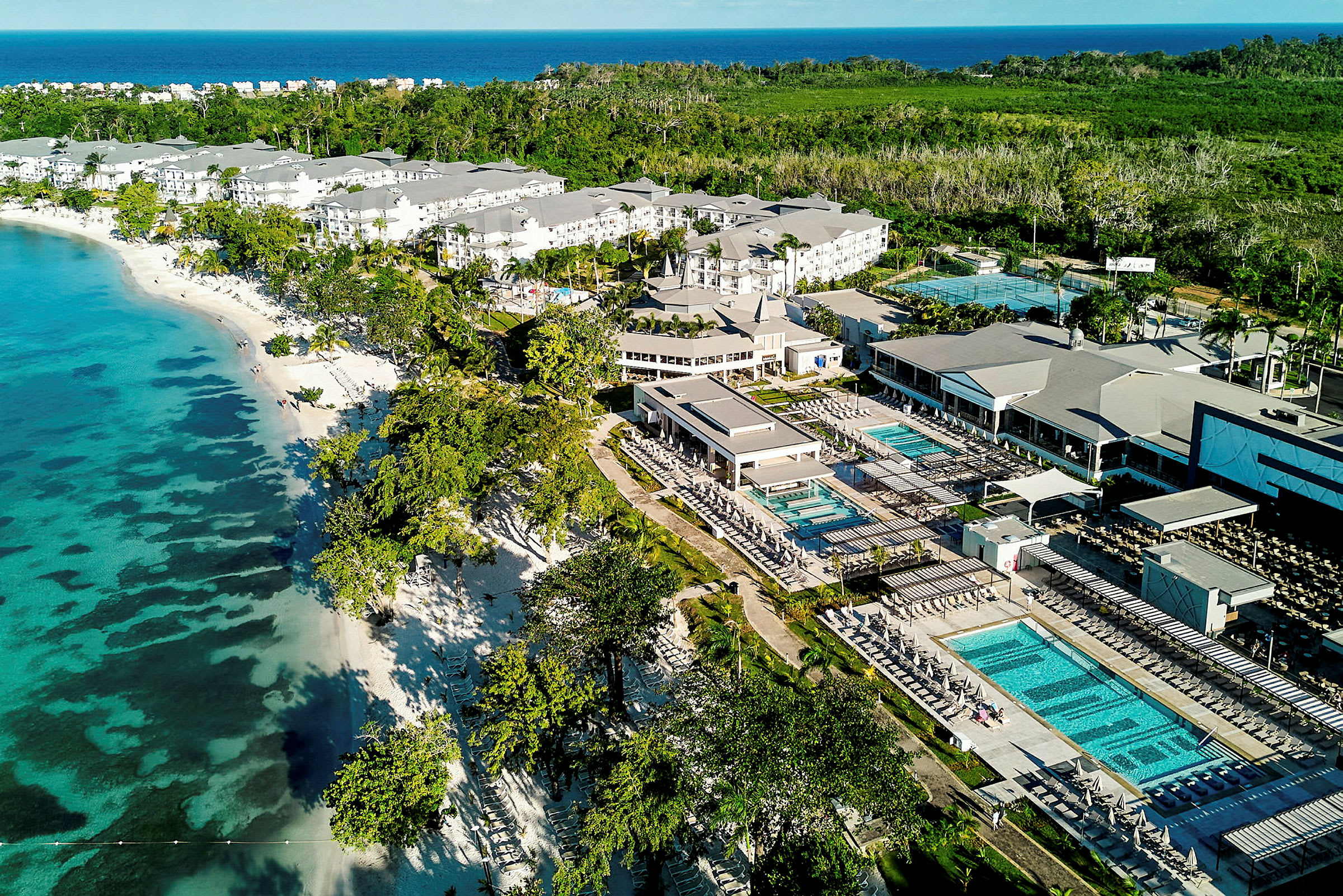 Luftfoto af hotelkompleks ved stranden med pools og omkringliggende grønt område. (AI-genereret tekst)