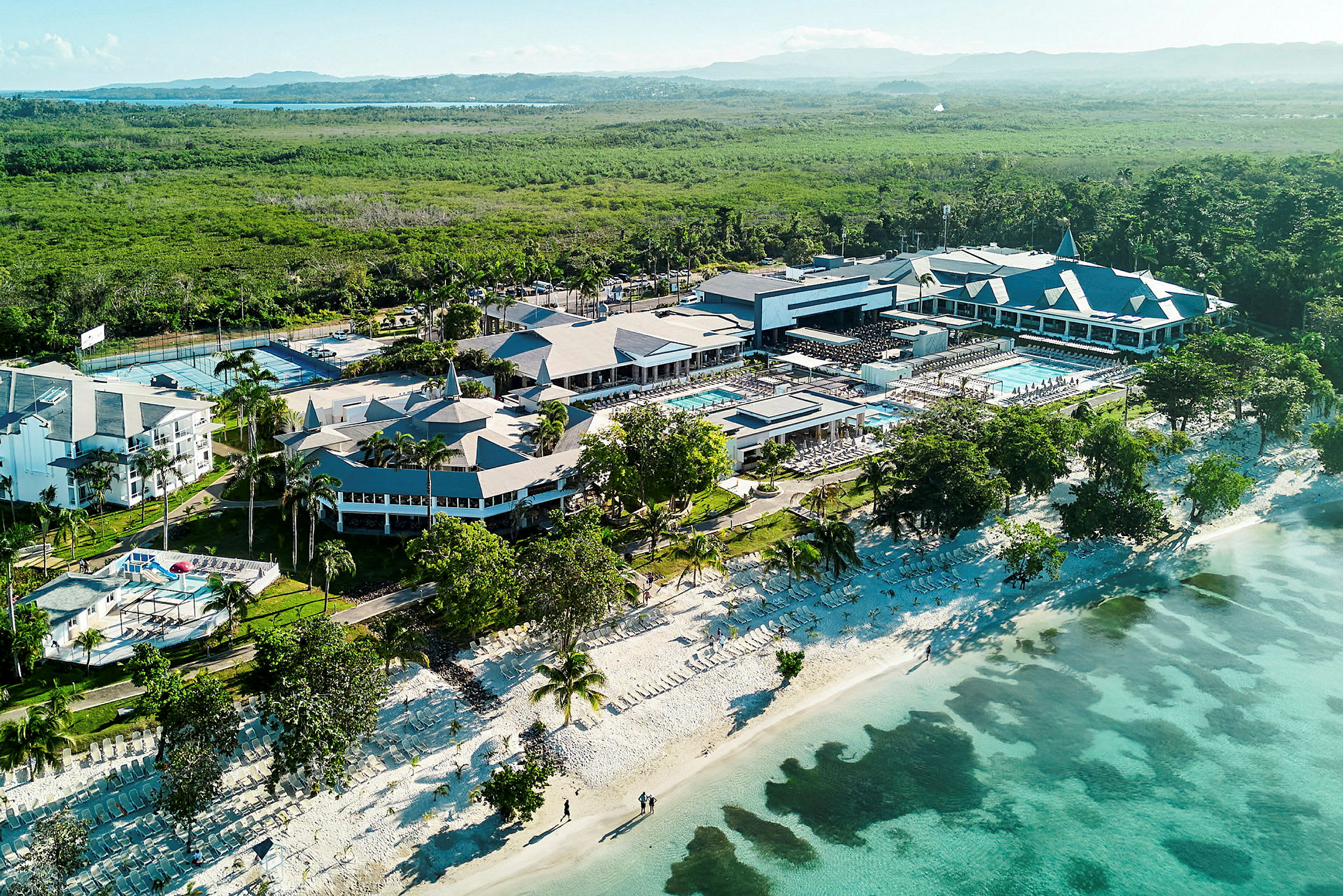 Luftfoto af en resort ved stranden med poolområder og omkringliggende grønne arealer. (AI-genereret tekst)