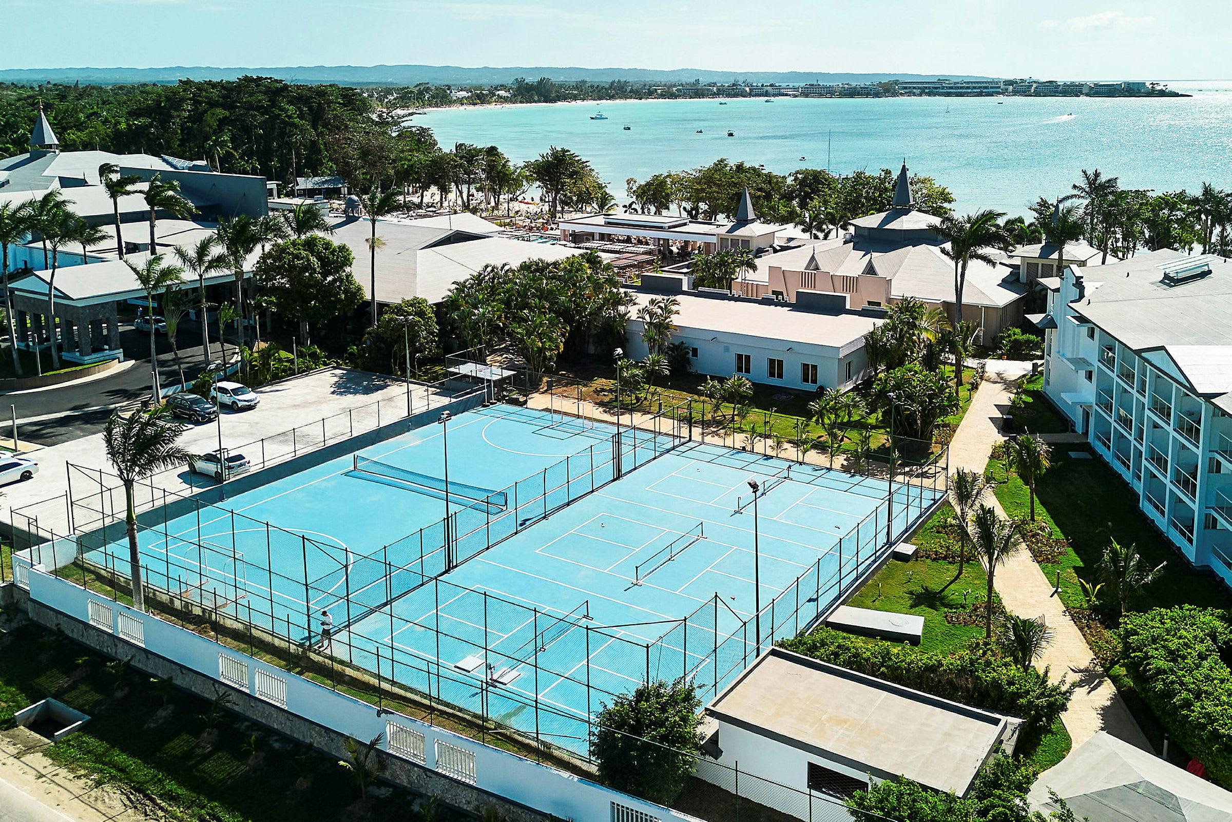 Udsigt over en tennisbane med blå belægning omgivet af bygninger og grønne områder tæt på havet. (AI-genereret tekst)