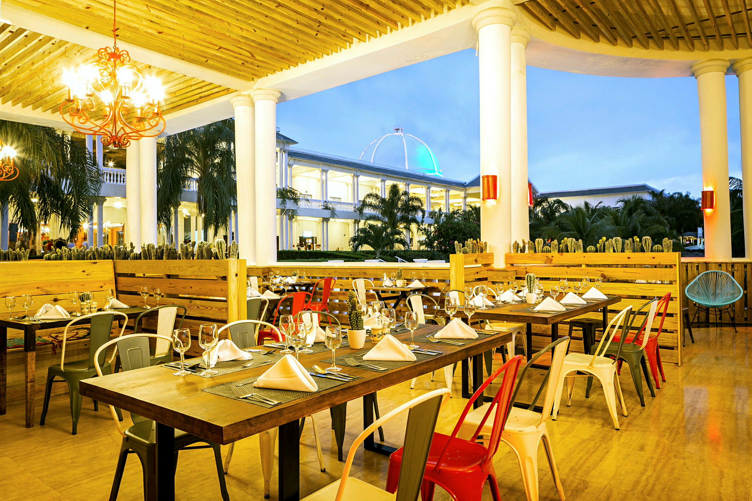 Billede af en restaurant med dækkede borde og farvede stole, beliggende under en træpergola med søjler. I baggrunden ses en bygning med en oplyst kuppelformet tag. (AI-genereret tekst)