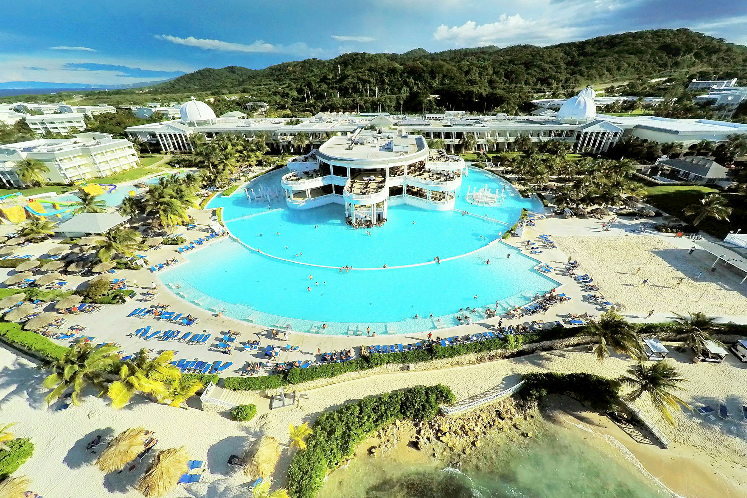 Et feriekompleks med en stor rund pool omgivet af solsenge og sandstrand, med grønne bjerge i baggrunden. (AI-genereret tekst)
