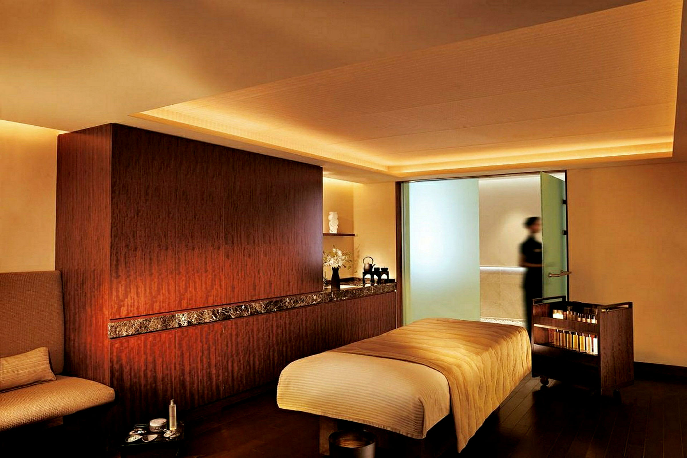Et spa-rum med en massagebriks dækket med beige stof og blød belysning. Et sidebord med produkter ses, og en person er synlig i baggrunden. (AI-genereret tekst)