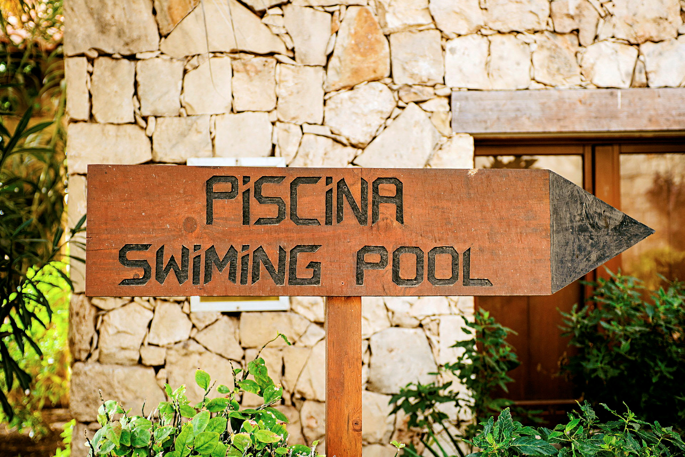 Skilt med teksten 'Piscina - Swimming Pool' foran stenmuren. (AI-genereret tekst)