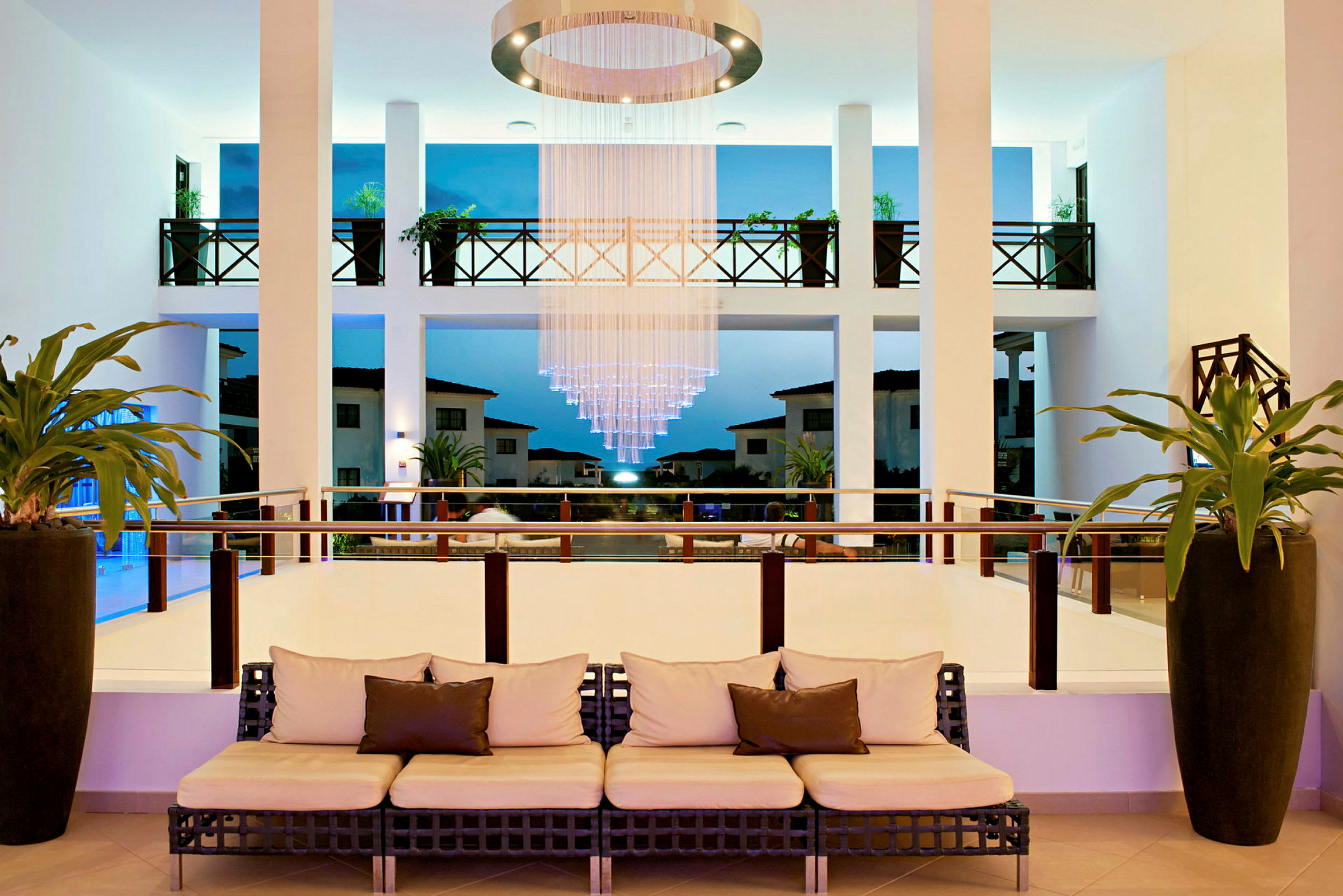En hotellobby med sofa, hængende lysekrone og udsigt til udendørsområdet. (AI-genereret tekst)