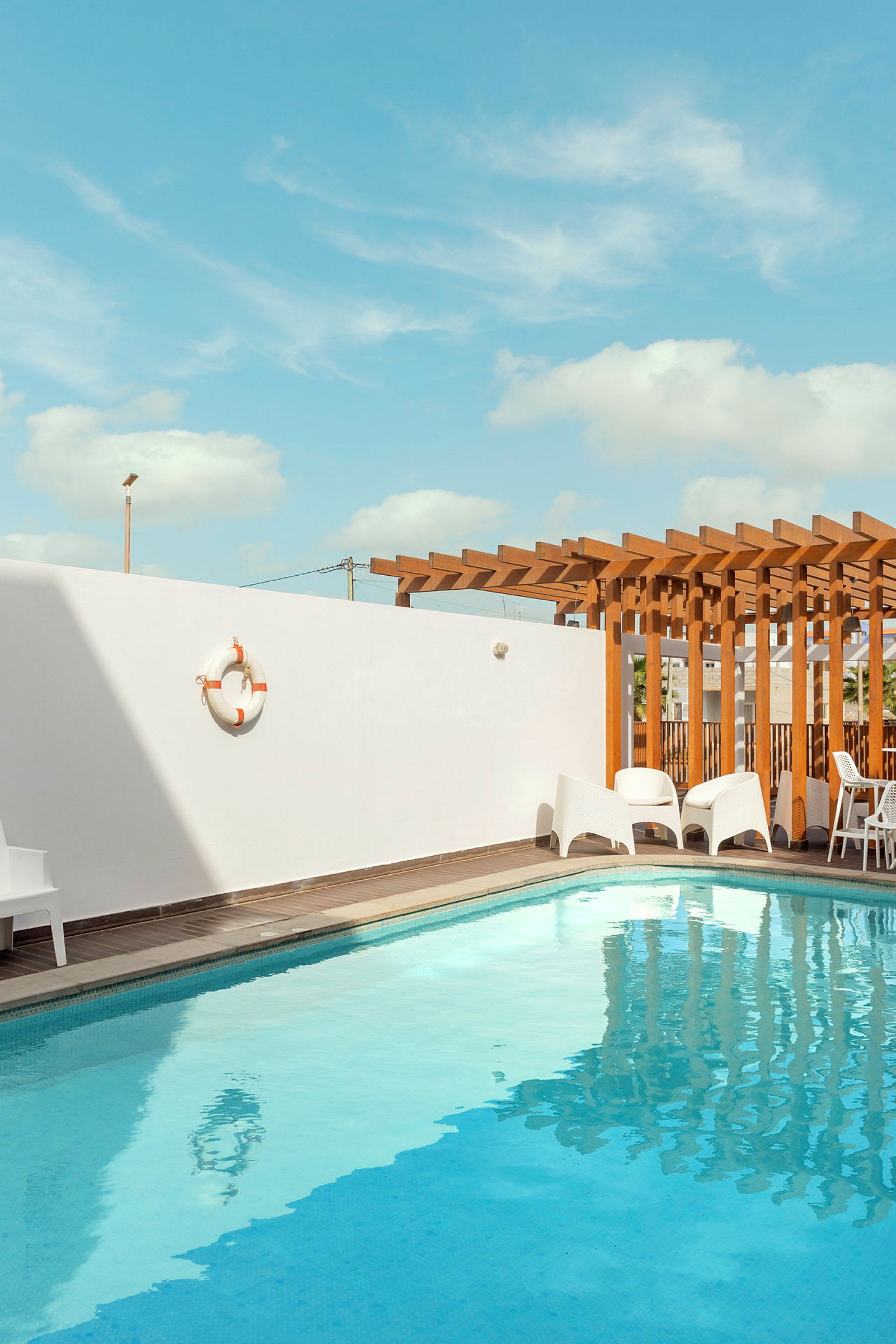 Udendørs swimmingpool med hvid væg og træpergola i baggrunden. (AI-genereret tekst)