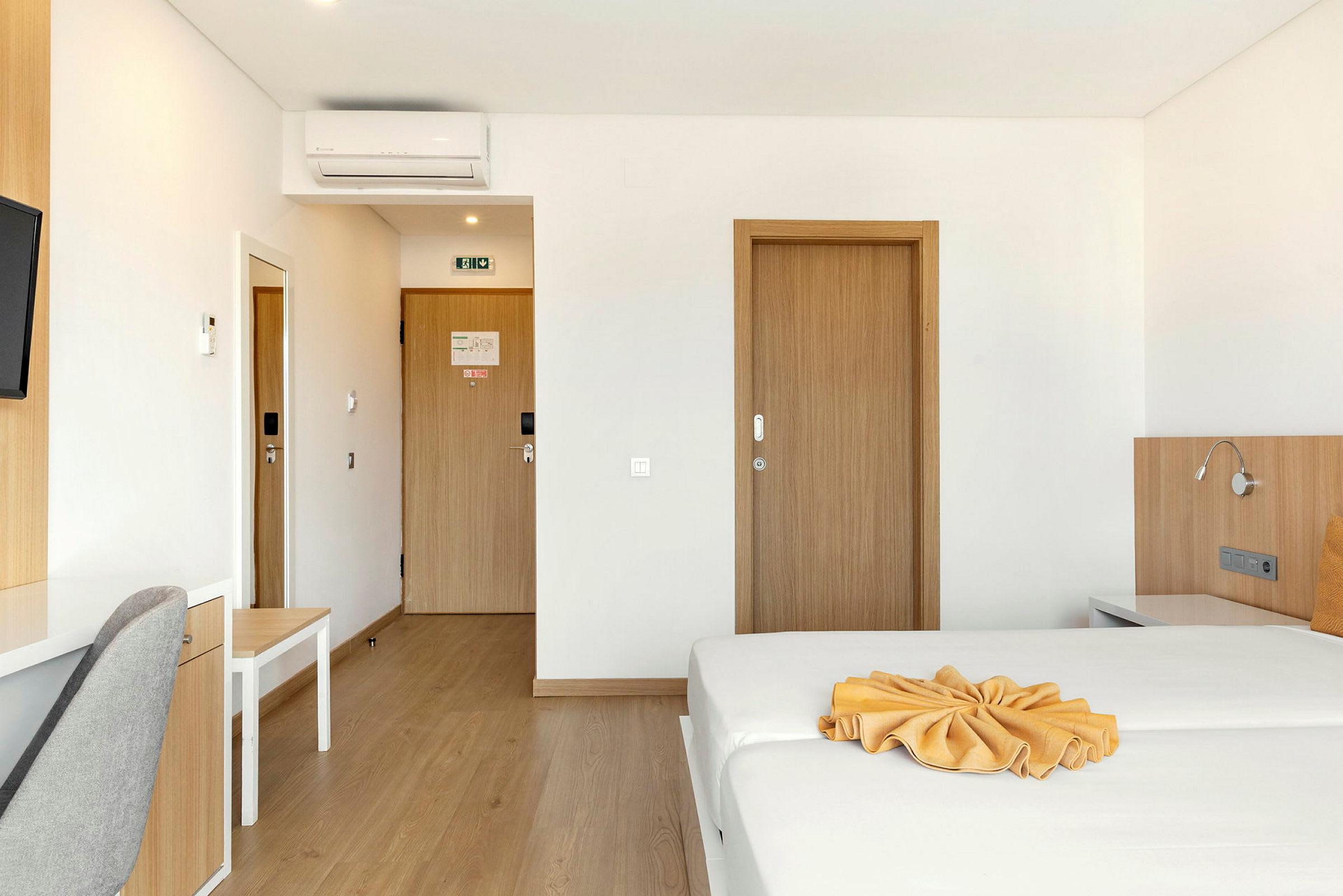 Et hotelværelse med en redt seng og et skrivebord ved siden af en grå stol, trædøre og en aircondition på væggen. (AI-genereret tekst)