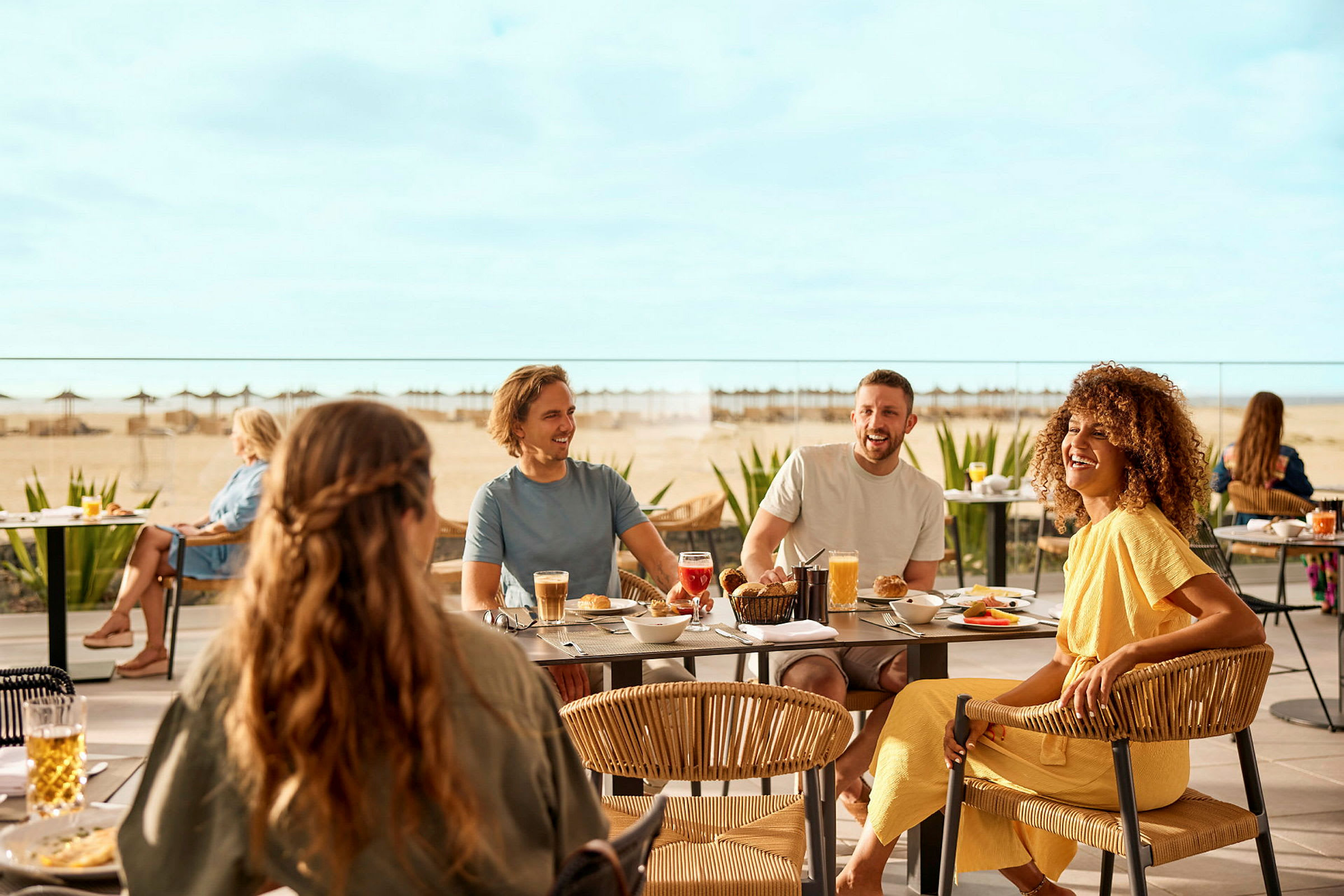 Personer sidder ved borde udendørs og nyder måltider, med en strand og havet i baggrunden. (AI-genereret tekst)