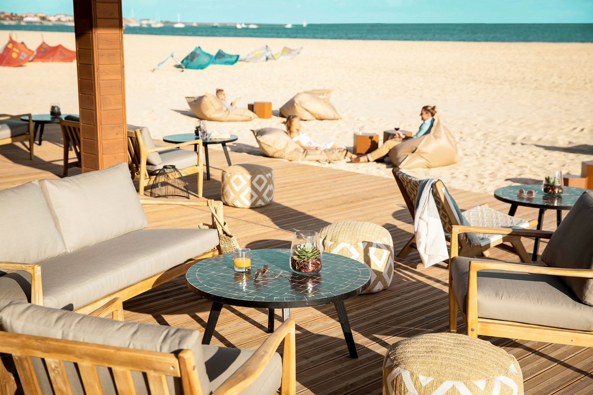 Terrasse med siddepladser og borde, i baggrunden en sandstrand og havet. (AI-genereret tekst)