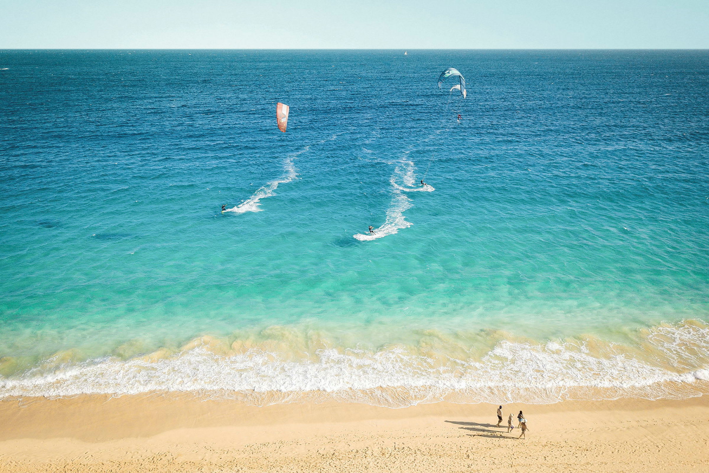 Strand med blåt hav, hvor personer kitesurfer og går tur. (AI-genereret tekst)