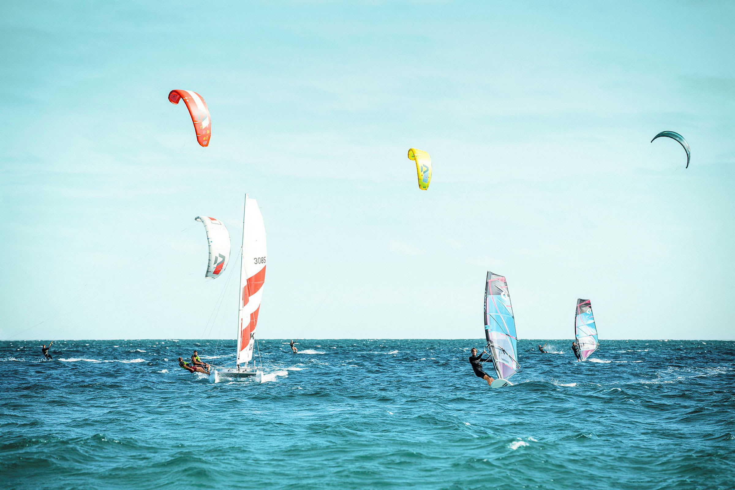 Flere personer deltager i forskellige vindsportsaktiviteter på havet, med sejlbåd, kitesurfing og windsurfingbrætter. (AI-genereret tekst)