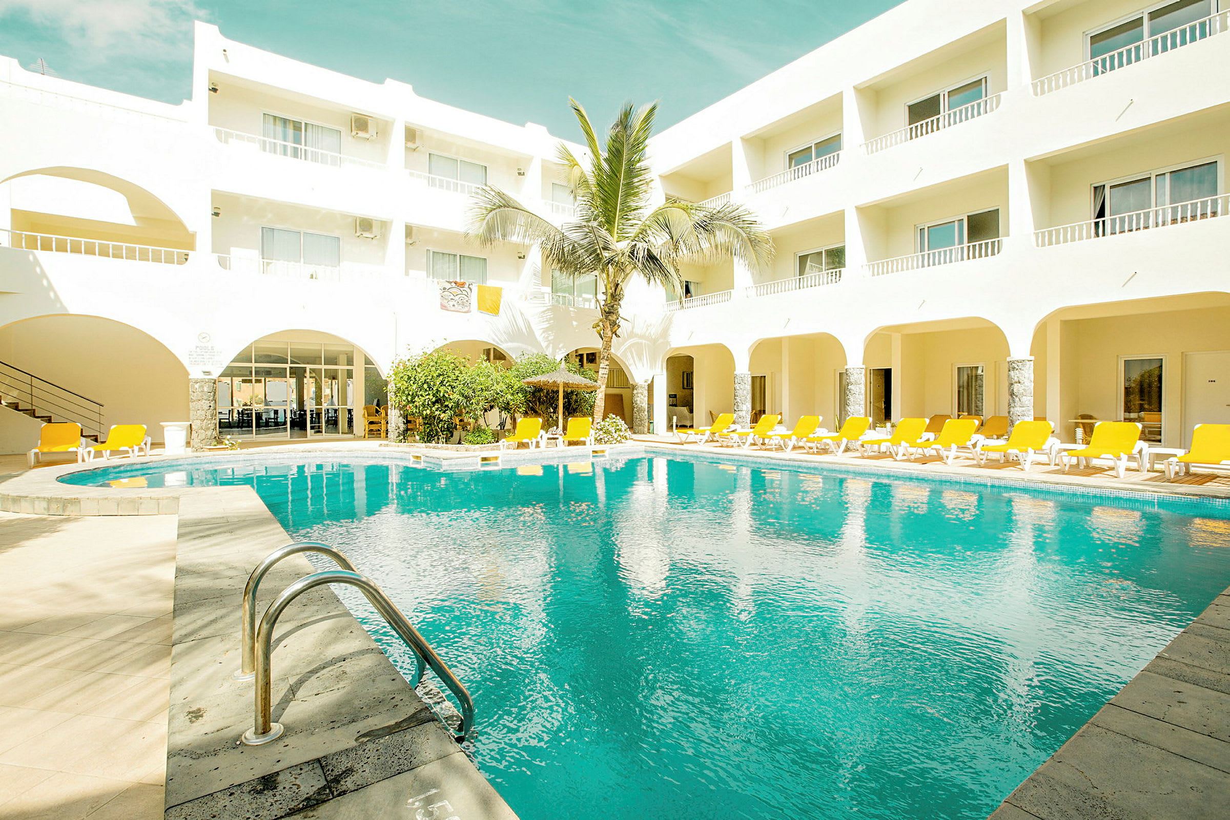 Hotelbygning med en pool og liggestole omkring. (AI-genereret tekst)