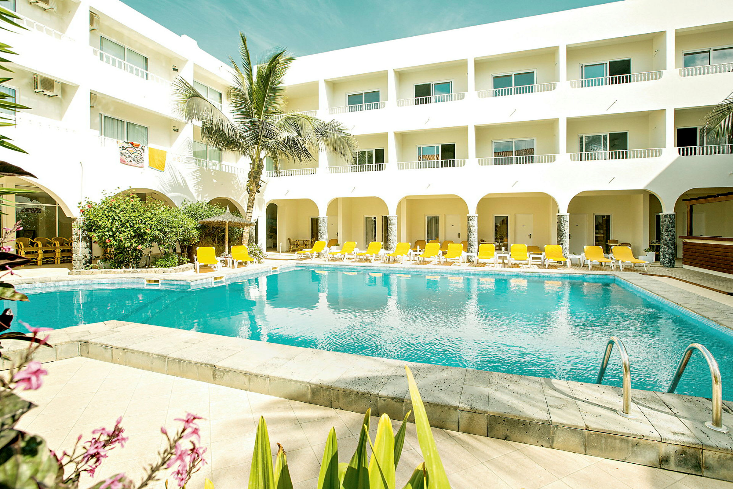 En udendørs pool med gule liggestole og en hvid bygning med altaner. (AI-genereret tekst)