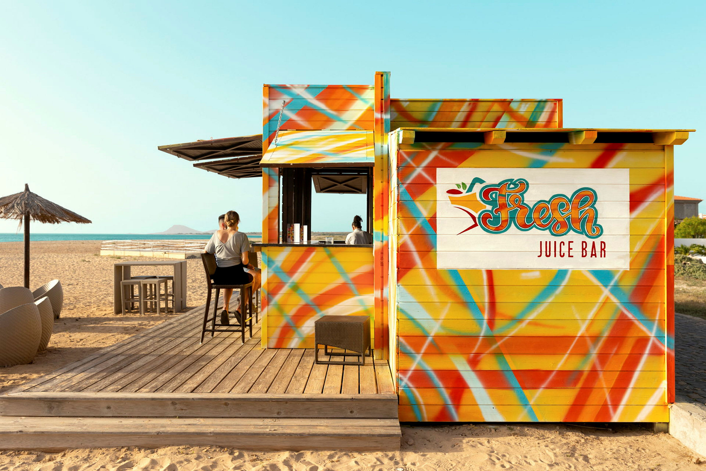 En farverig juicebar på stranden med personer, der sidder på barstole. (AI-genereret tekst)