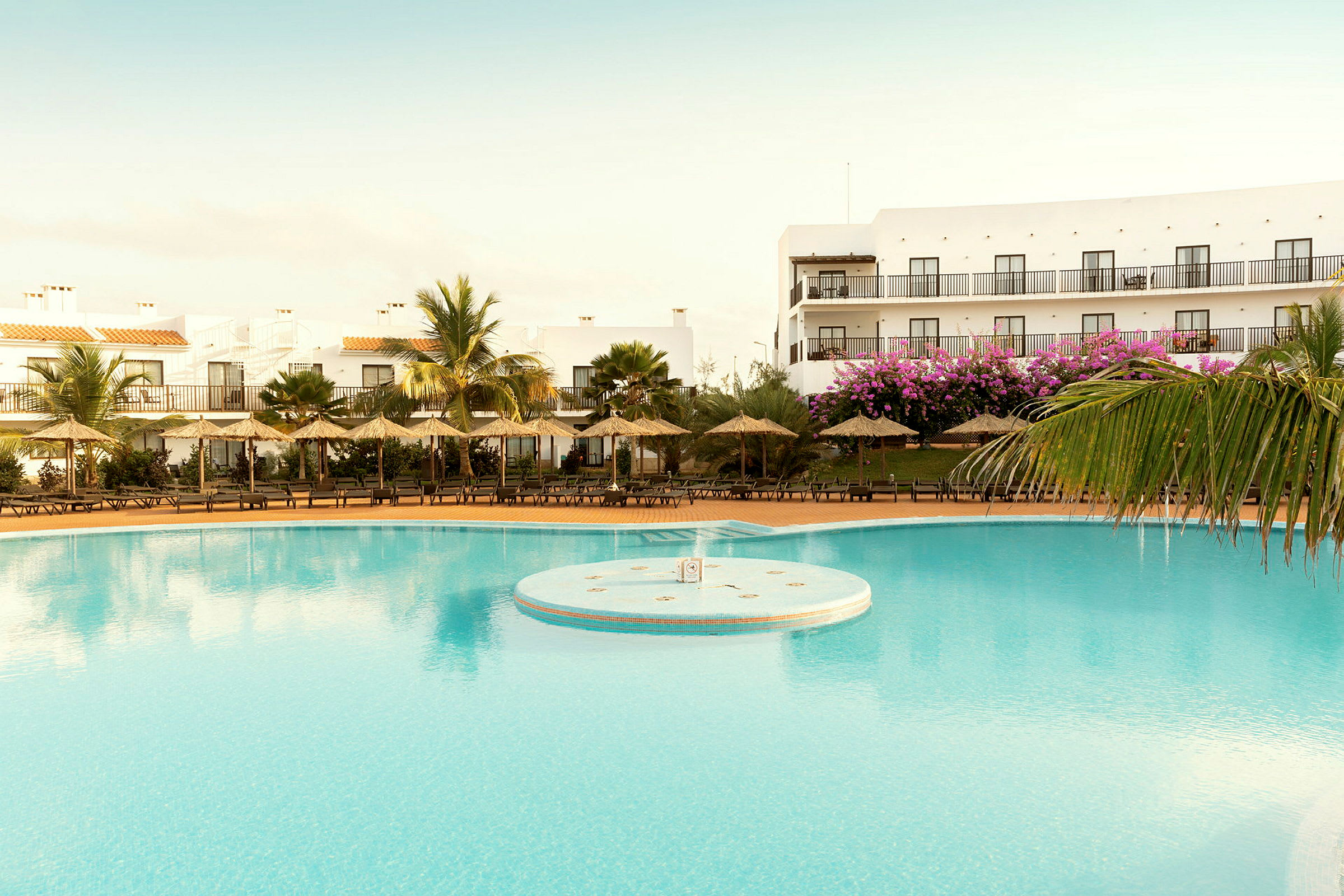 Billede af en pool ved en resort med parasoller og bygninger i baggrunden. (AI-genereret tekst)
