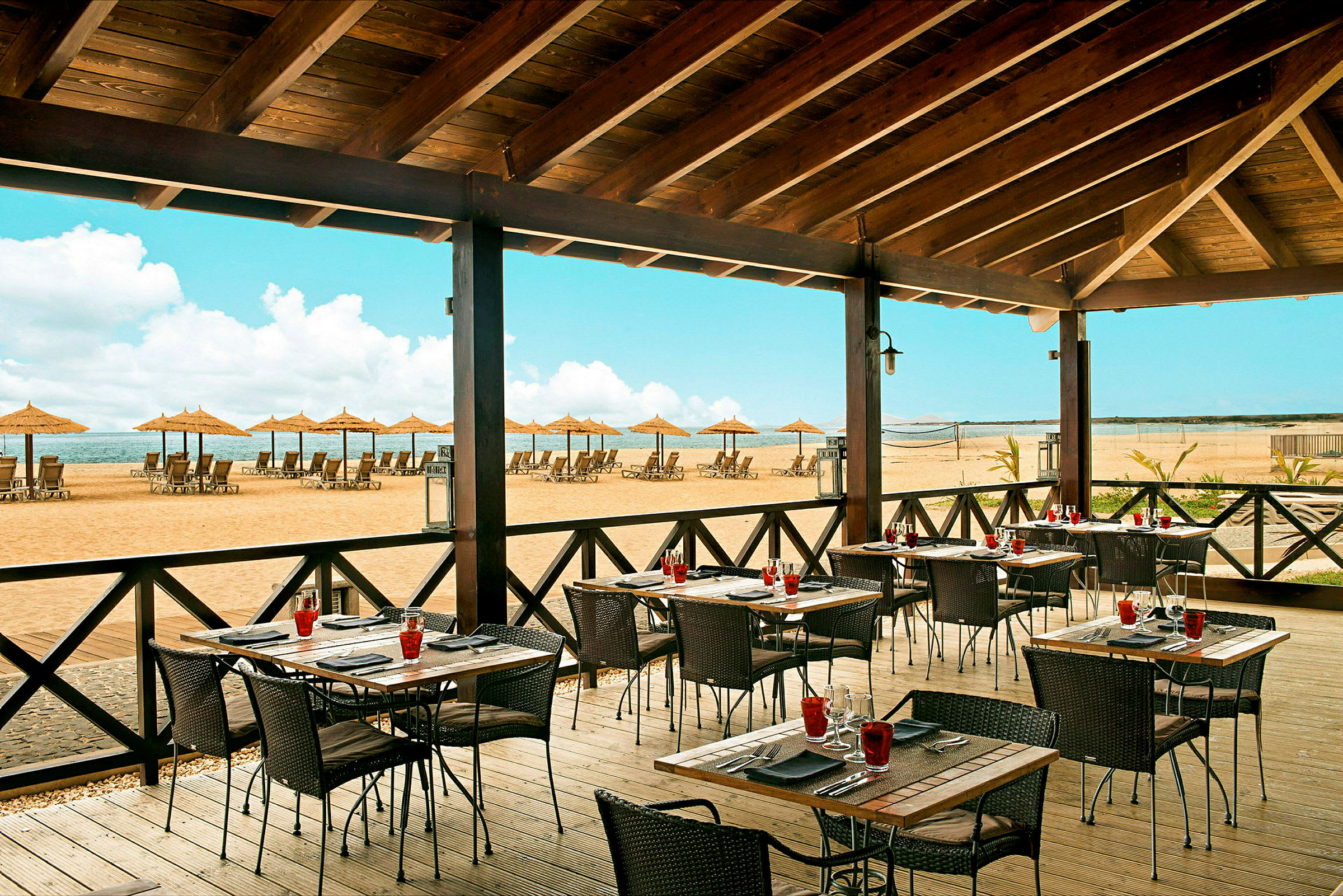 Restaurant med udendørs servering og udsigt over en strand med liggestole og parasoller. (AI-genereret tekst)