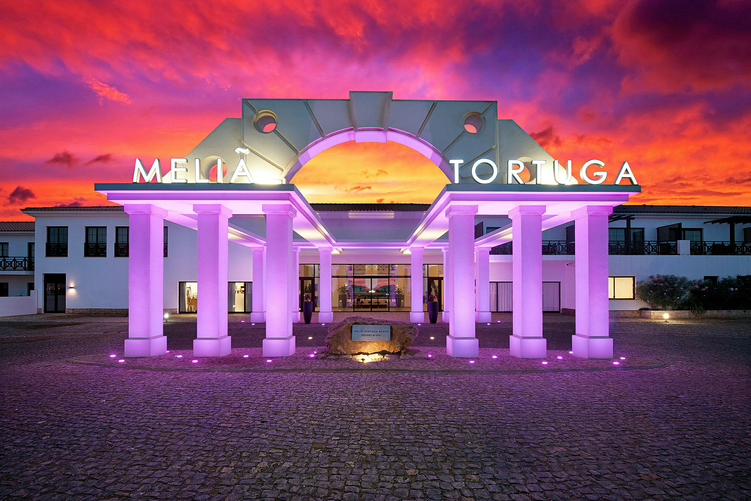 Indgangen til hotellet med belyste søjler og en farverig solnedgang i baggrunden. (AI-genereret tekst)