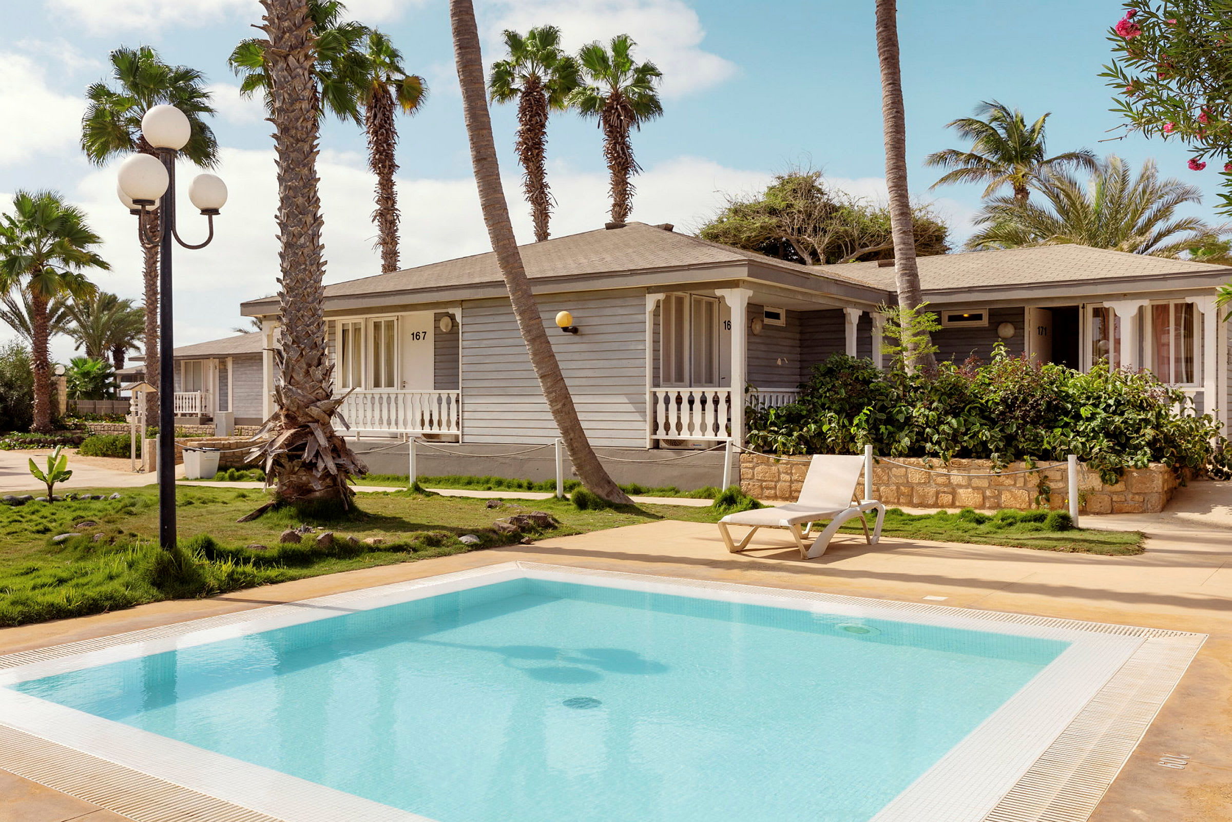 En udendørs pool med en solseng, omgivet af grønt og palmer samt huse i baggrunden. (AI-genereret tekst)