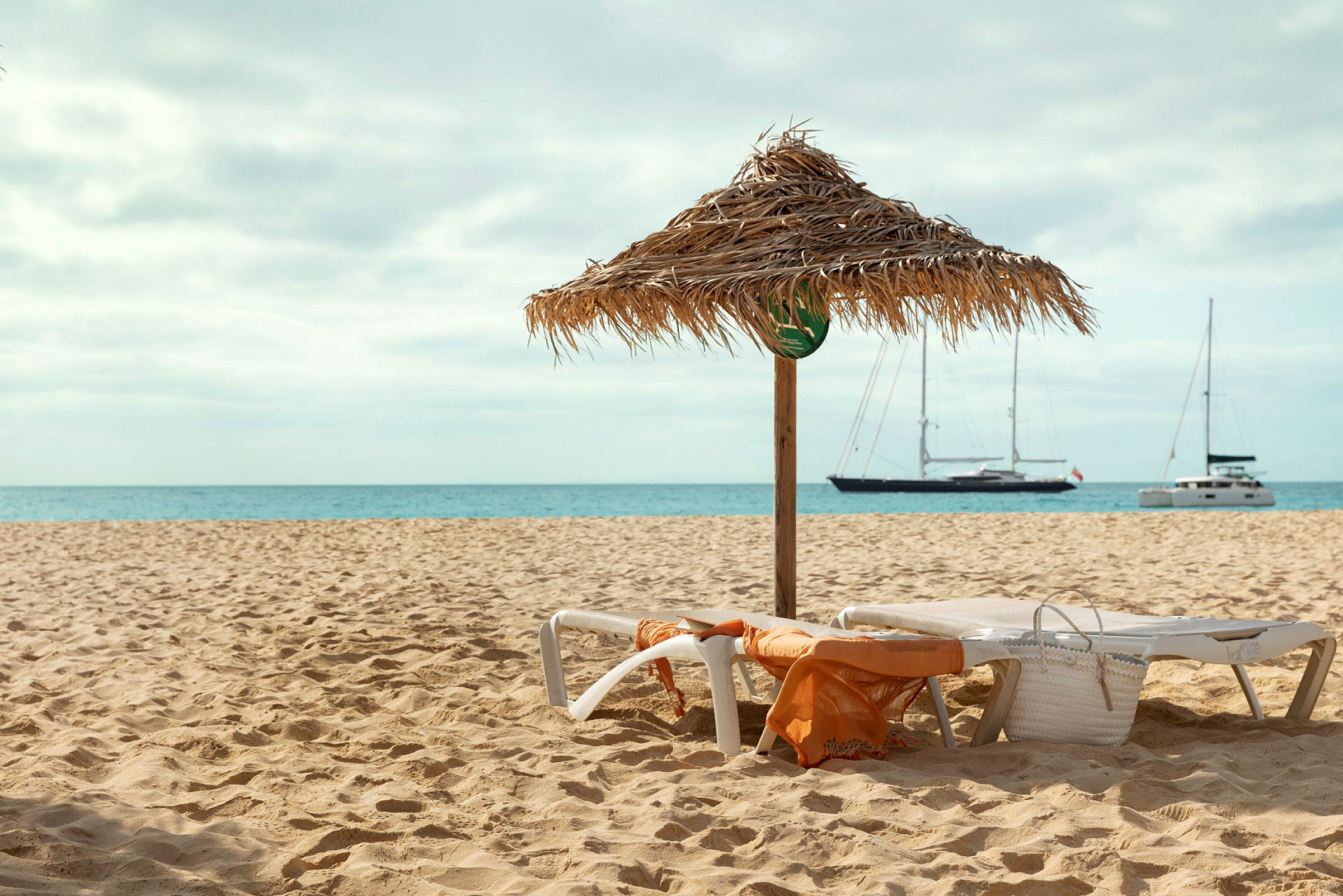 Strand med parasol og liggestole. Sejlskibe ses på havet. (AI-genereret tekst)