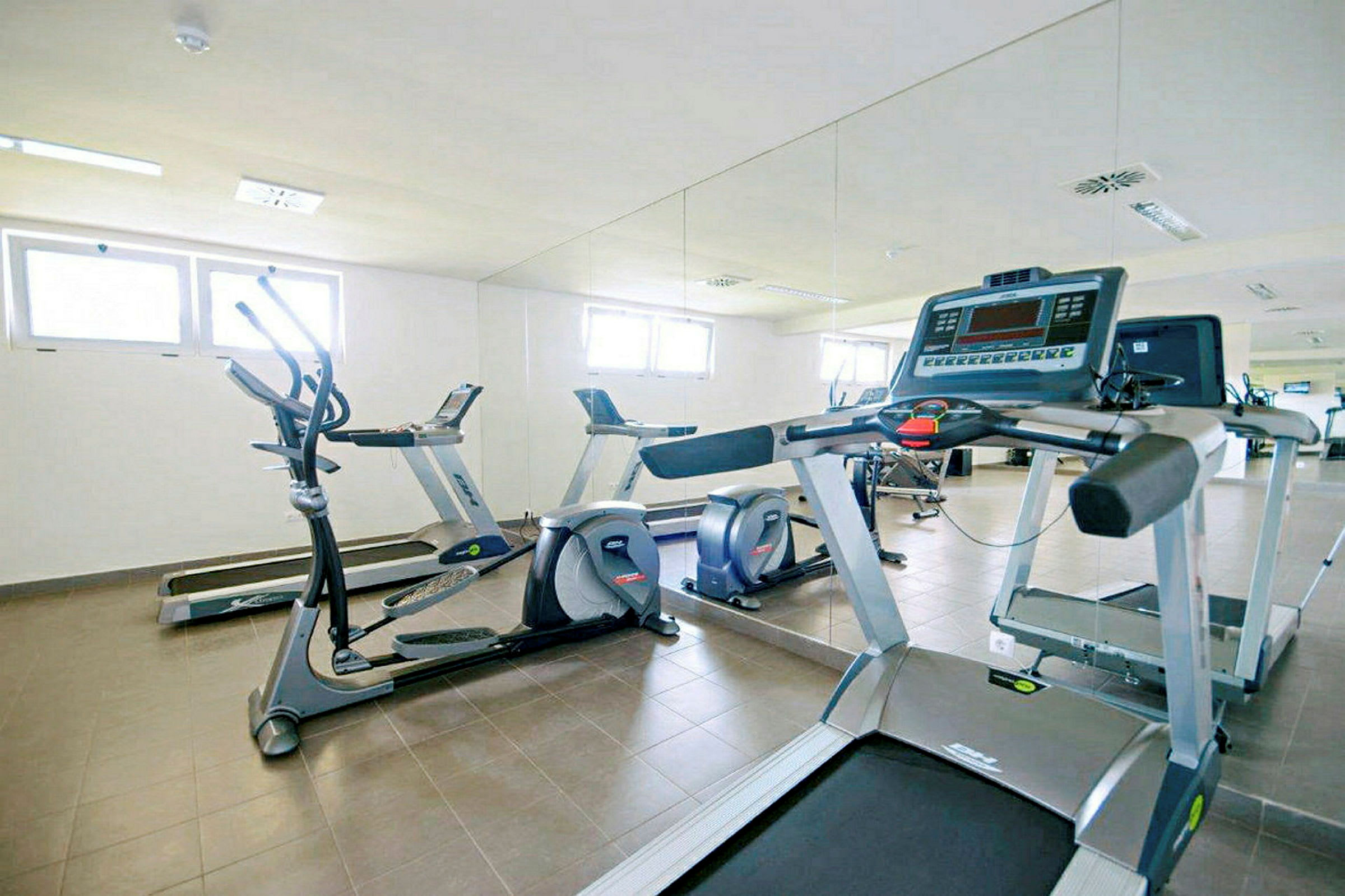 Billede af et fitnesscenter med træningsudstyr, herunder løbebånd og elliptiske maskiner foran et spejl. (AI-genereret tekst)