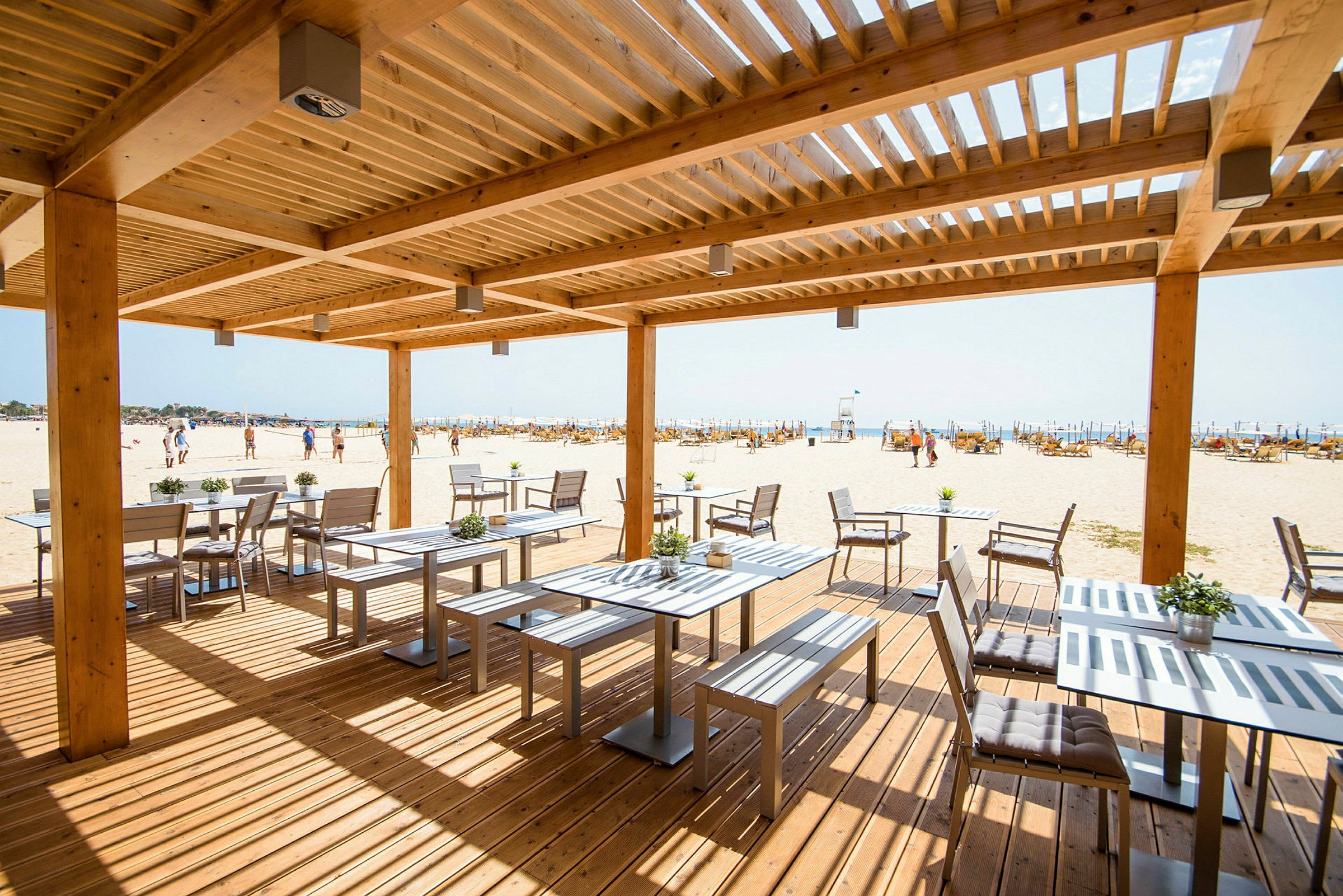 En træpergola over udendørs servering med udsigt over en sandstrand. (AI-genereret tekst)