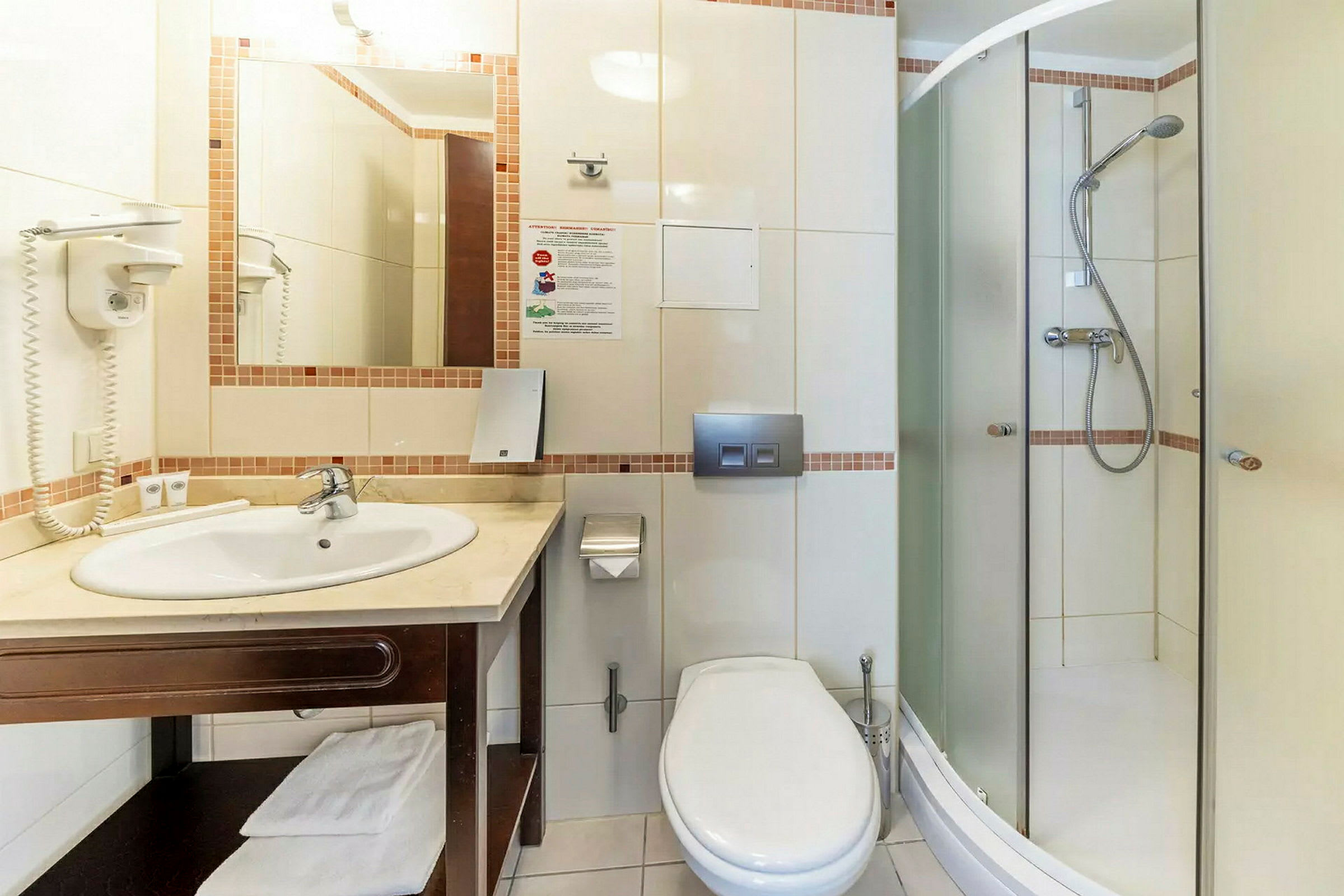 Hotelbadeværelse med brusekabine, toilet, håndvask og spejl. (AI-genereret tekst)