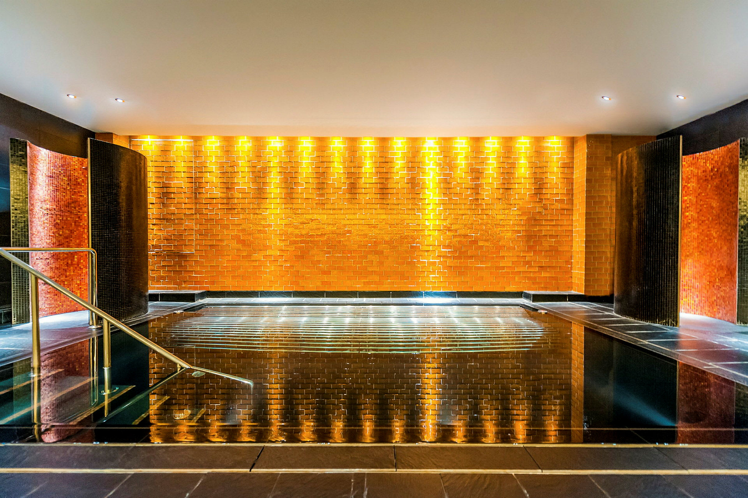Et indendørsspa med et skinnende sort flisebelagt poolområde, omgivet af en væg med orange fliser og belysning. (AI-genereret tekst)