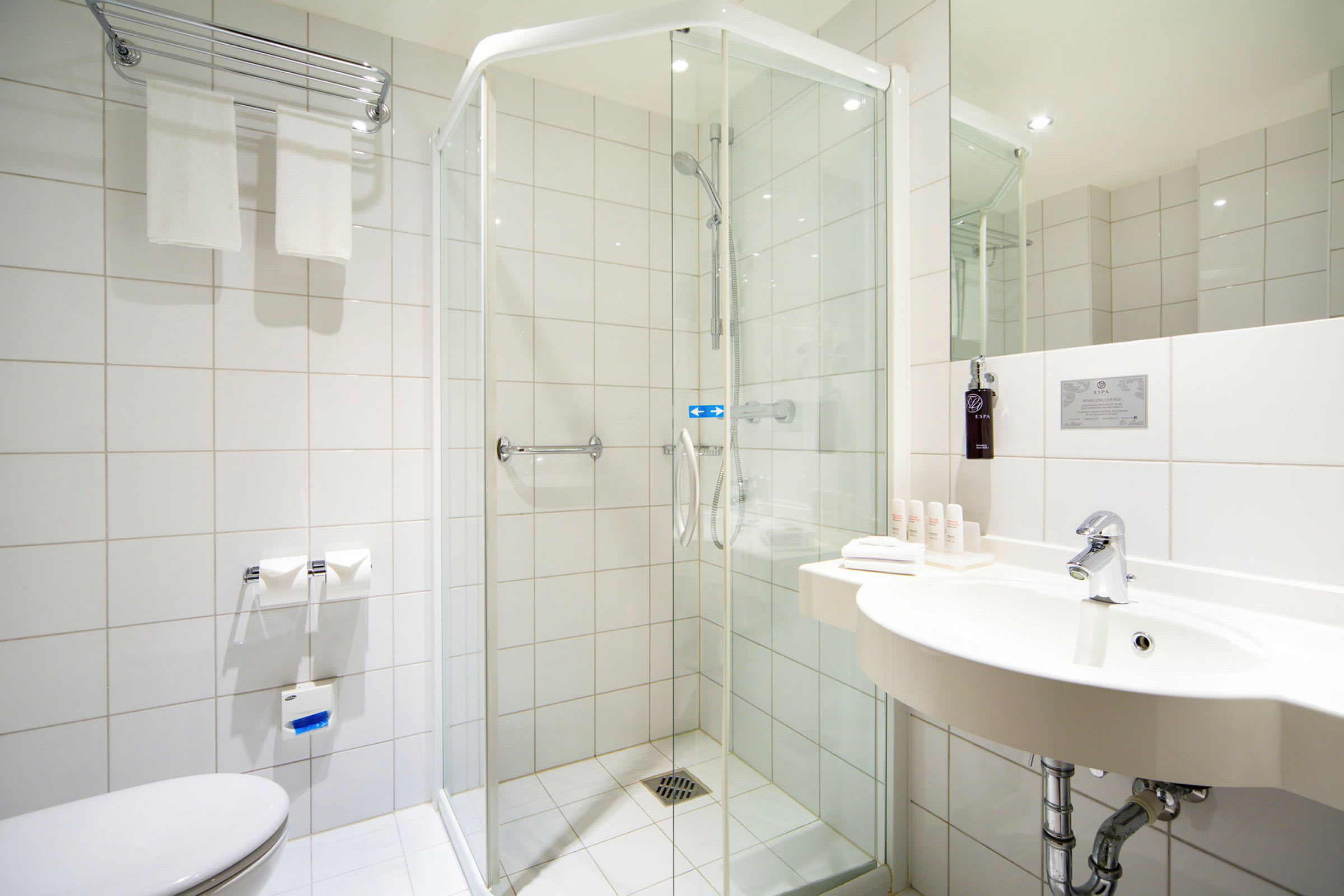 Et moderne badeværelse med brusekabine, håndvask og toilet. Håndklæder og toiletartikler er synlige, og interiøret er lyst med hvide flisevægge. (AI-genereret tekst)