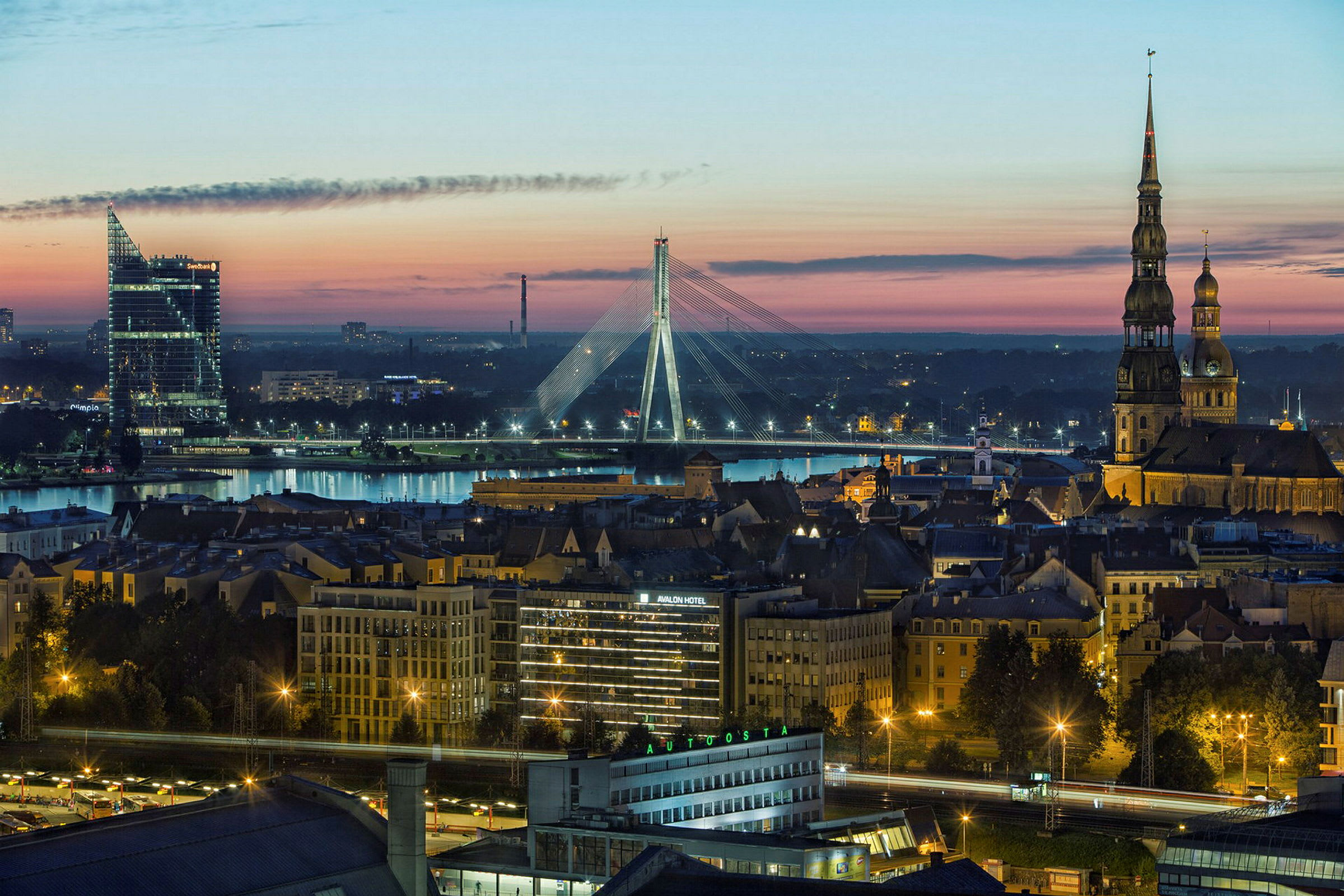 Panoramabillede af en by i skumringen, med en flod, oplyste bygninger og en bro i baggrunden. (AI-genereret tekst)