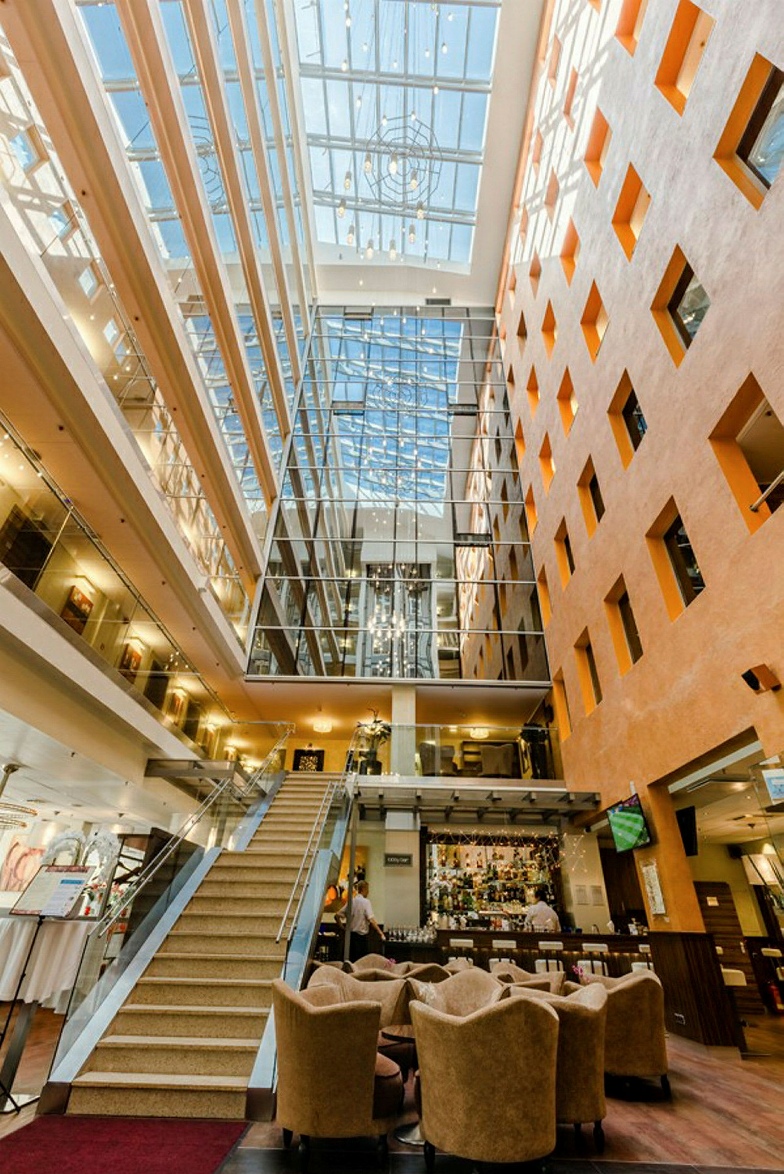 Billede af en lys hotellobby med en trappe, siddegrupper og en reception dekorativt belyst af naturligt lys fra et ovenlysvindue. (AI-genereret tekst)