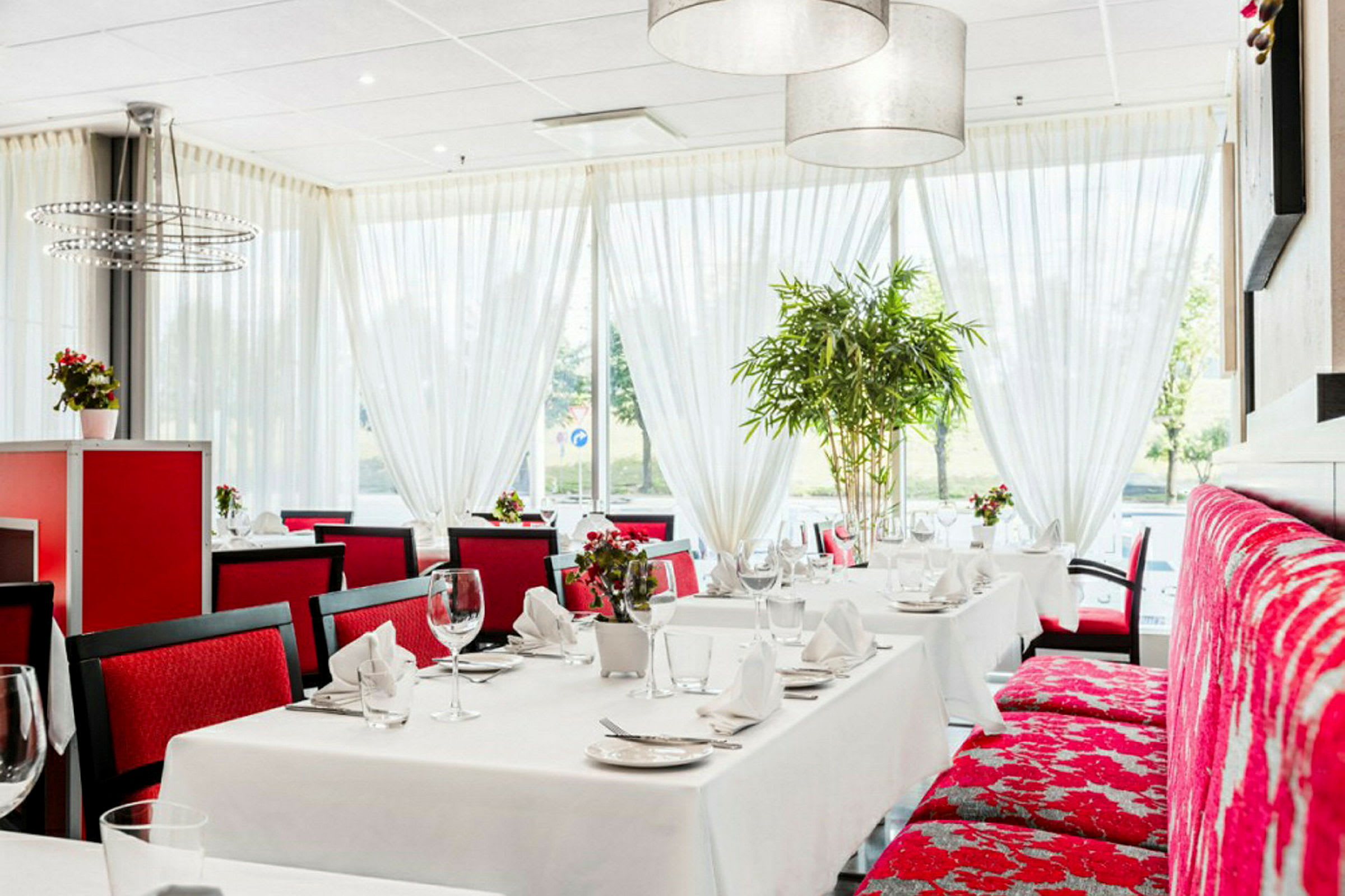 En restaurant med hvide duge og røde stole, dekoreret med store vinduer og grønne planter. (AI-genereret tekst)