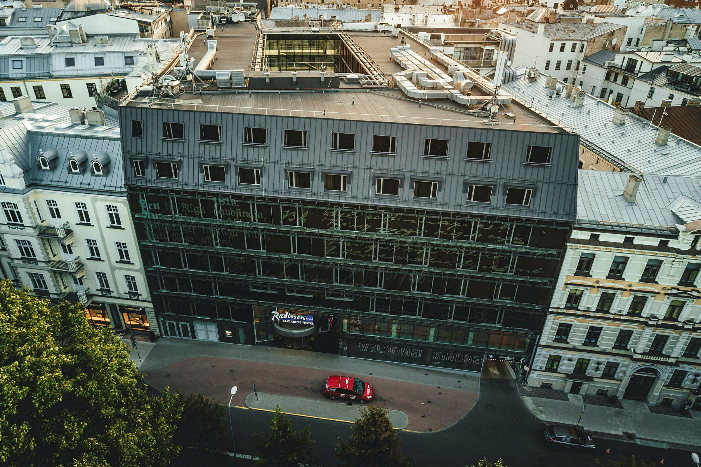 Luftfoto af en bygning med glasfacade og flere etager omgivet af andre bygninger. (AI-genereret tekst)
