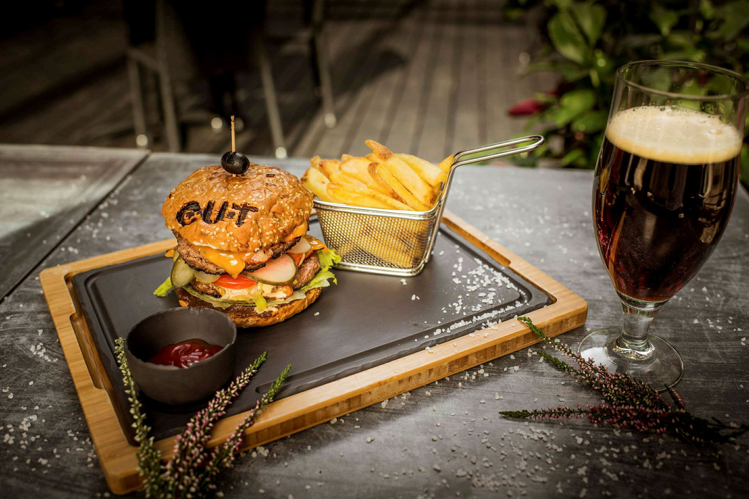 En tallerken med en burger og pommes frites samt en skål med ketchup, placeret på et bord ved siden af et glas mørk drik. (AI-genereret tekst)