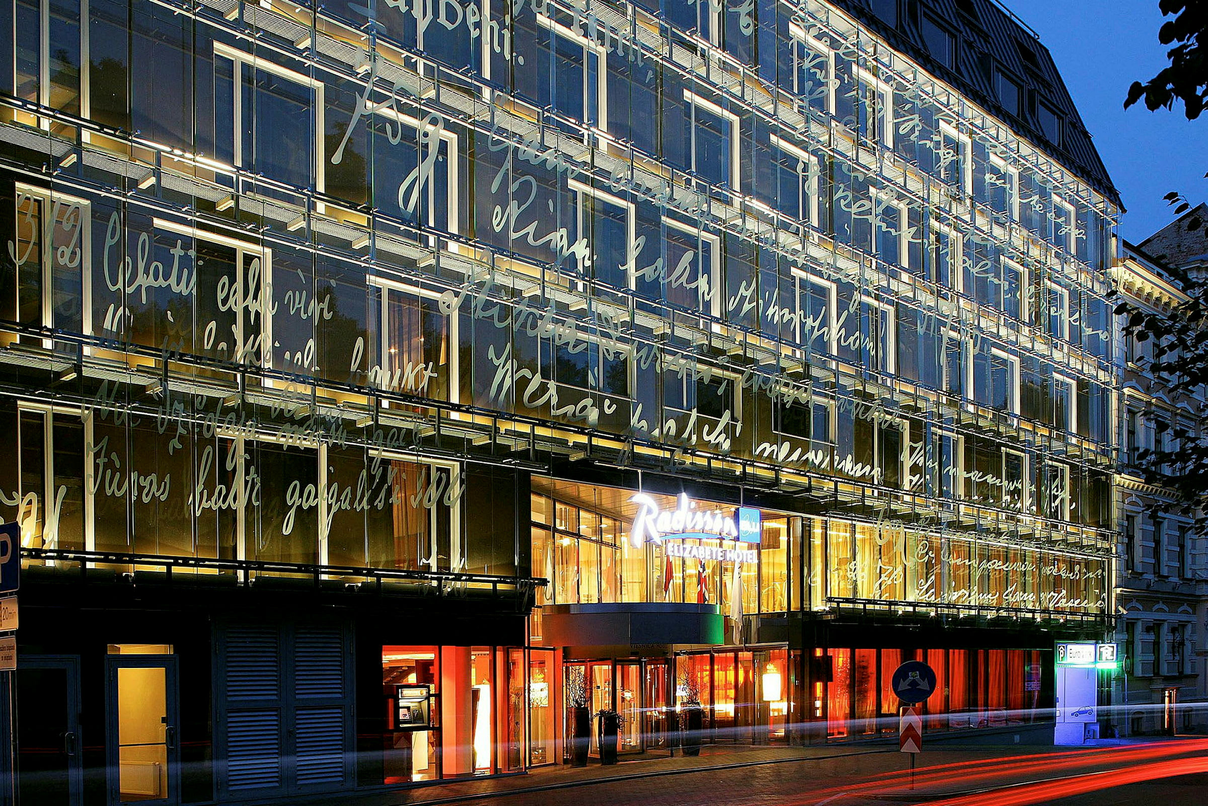En moderne bygning med glasfacade oplyst om aftenen, dekoreret med skrifttyper. Indgangen til en hotelkæde er tydeligt synlig. (AI-genereret tekst)