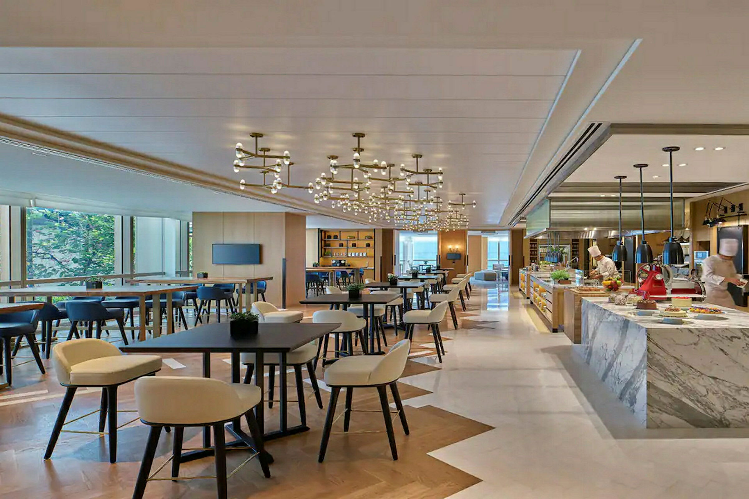 En moderne restaurant med borde, stole og et køkkenområde, hvor to personer arbejder. (AI-genereret tekst)