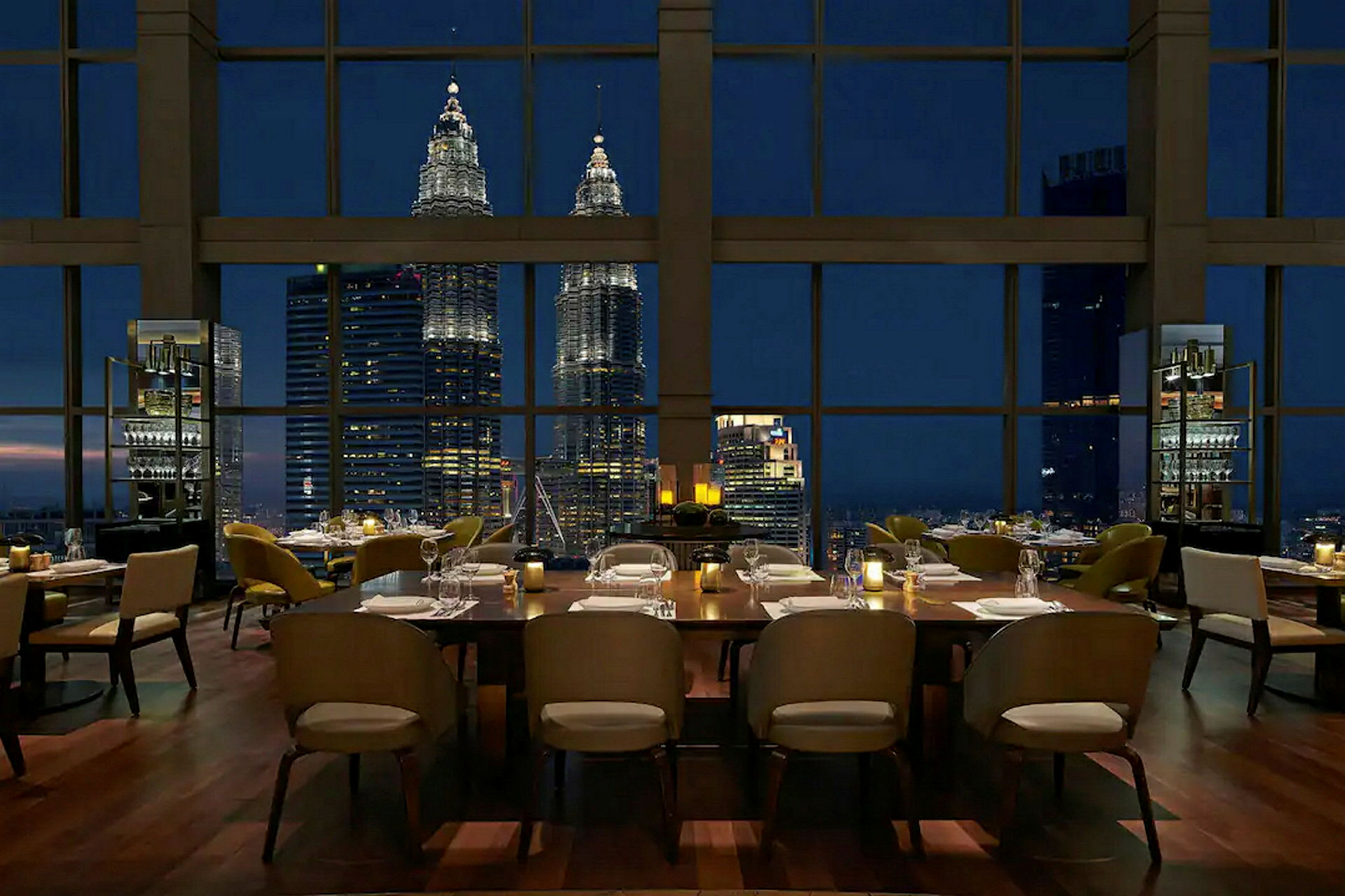 En restaurant med dækkede borde og udsigt til Petronas Twin Towers gennem store vinduer om aftenen. (AI-genereret tekst)