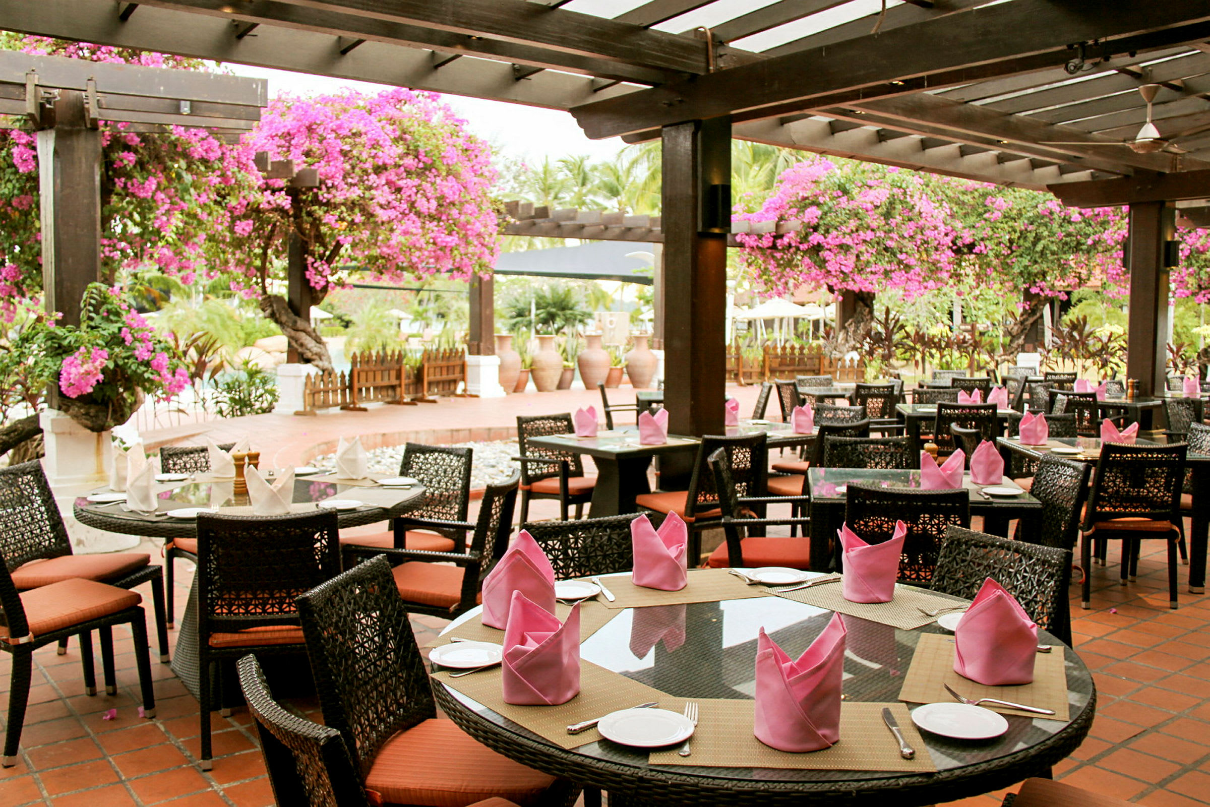 En udendørs restaurant med borde og stole under en pergola dekoreret med lyserøde blomster. (AI-genereret tekst)