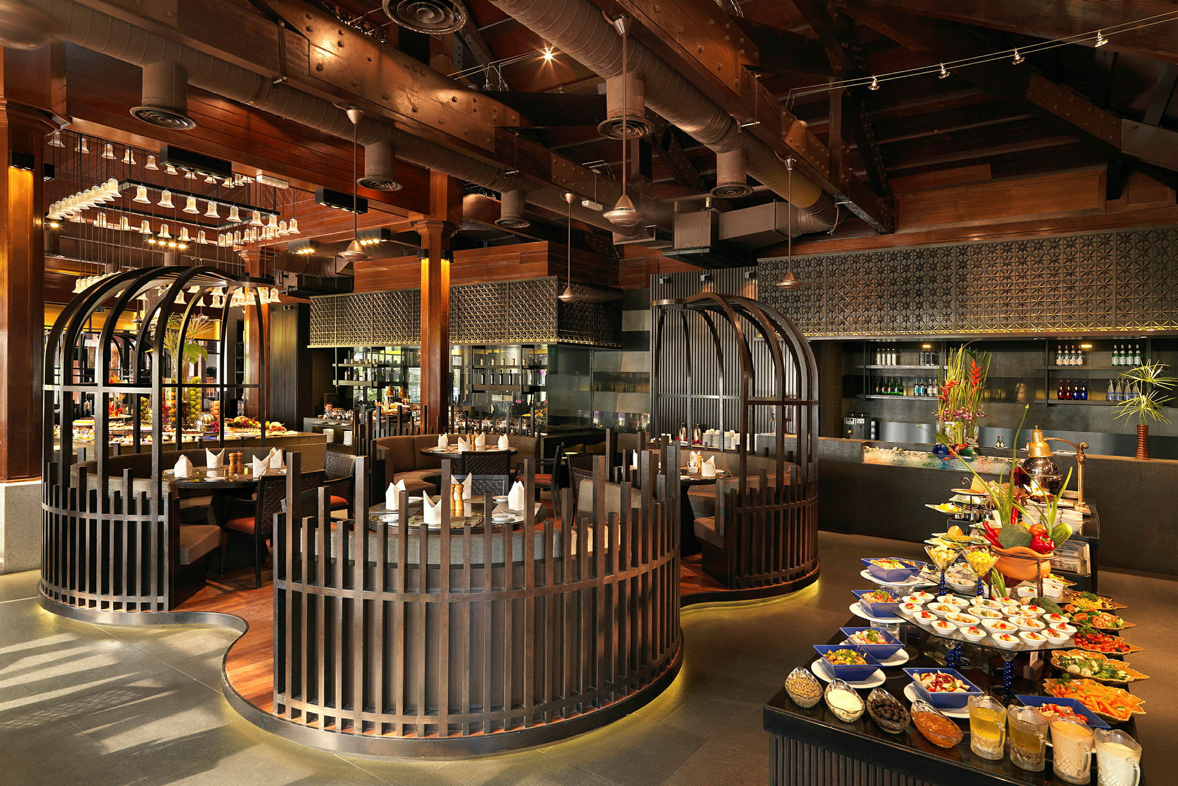 En restaurant med elegant interiør, herunder siddeområder omgivet af træafskærmninger, en madbuffet med farverige retter og drikkevarer samt en moderne atmosfære med synlige loftstrukturer. (AI-genereret tekst)