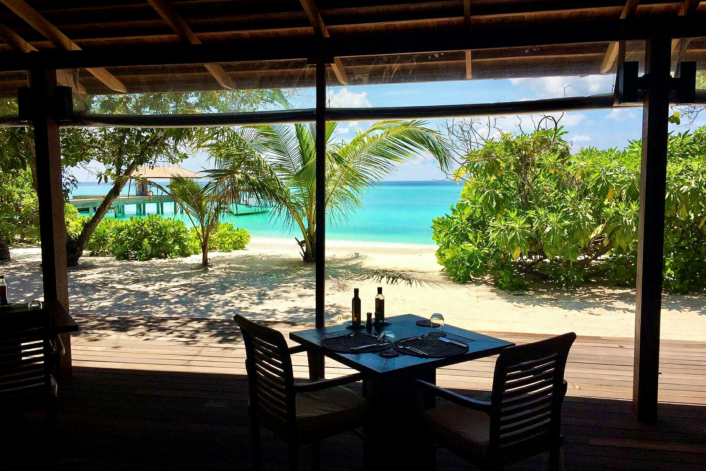 Udsigt fra en restaurant mod en tropisk strand med turkis hav og frodig vegetation. (AI-genereret tekst)