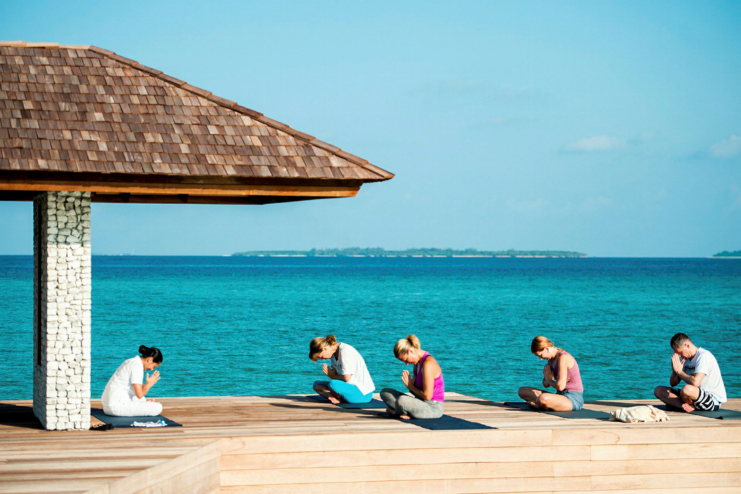 En gruppe mennesker udfører yogaøvelser ved havet under et træoverdækning. (AI-genereret tekst)