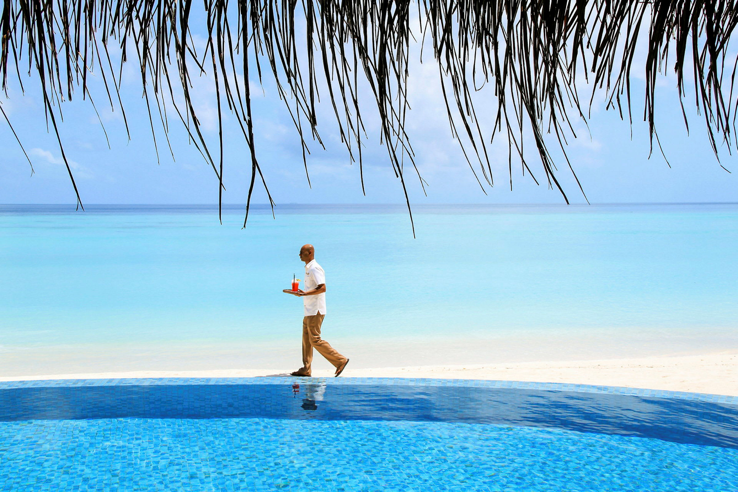 En person går med en bakke med en drink tæt ved en klarblå pool ved stranden. (AI-genereret tekst)