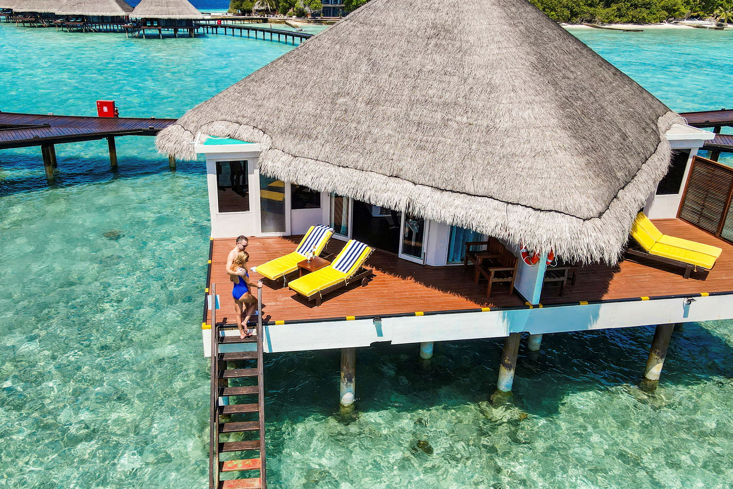 Bungalow på pæle over klart turkis vand med solterrasse og strandstole. (AI-genereret tekst)