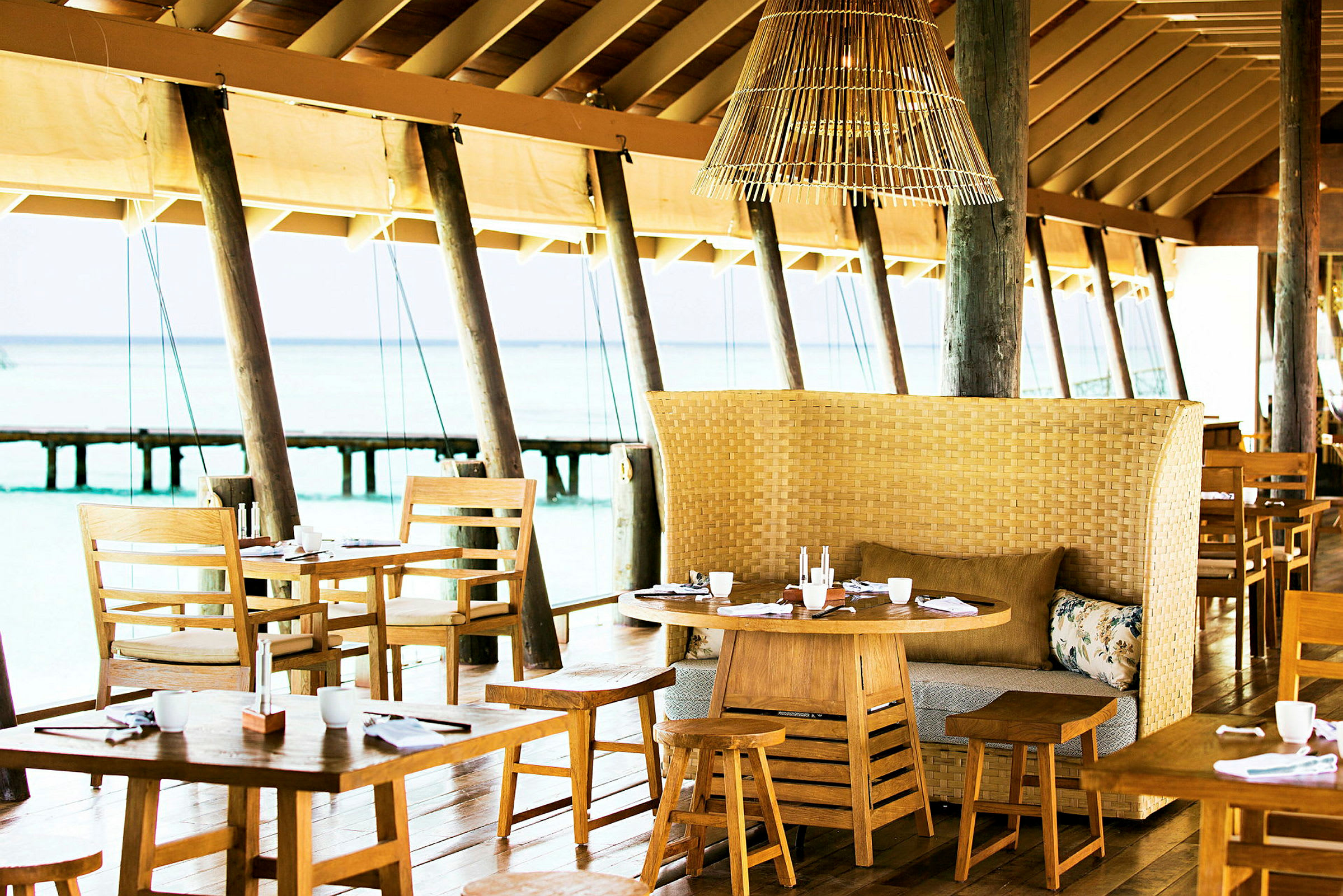 En restaurant med træborde og stole samt en flettet bænk med puder, beliggende ved havet. (AI-genereret tekst)