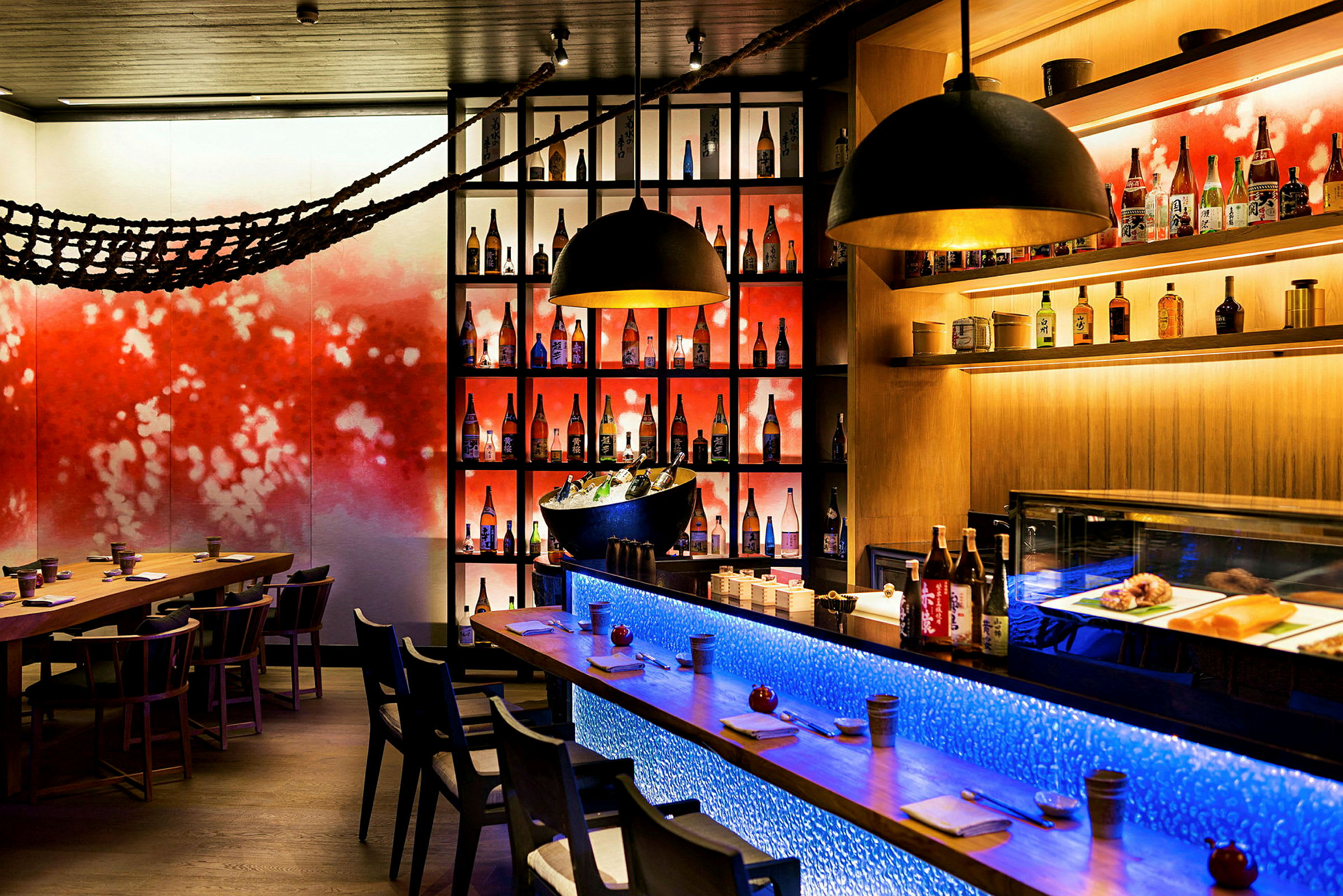 En restaurantinteriør med et bord, stole og en bardisk, samt hylder med flasker og en farverig vægdekoration. (AI-genereret tekst)