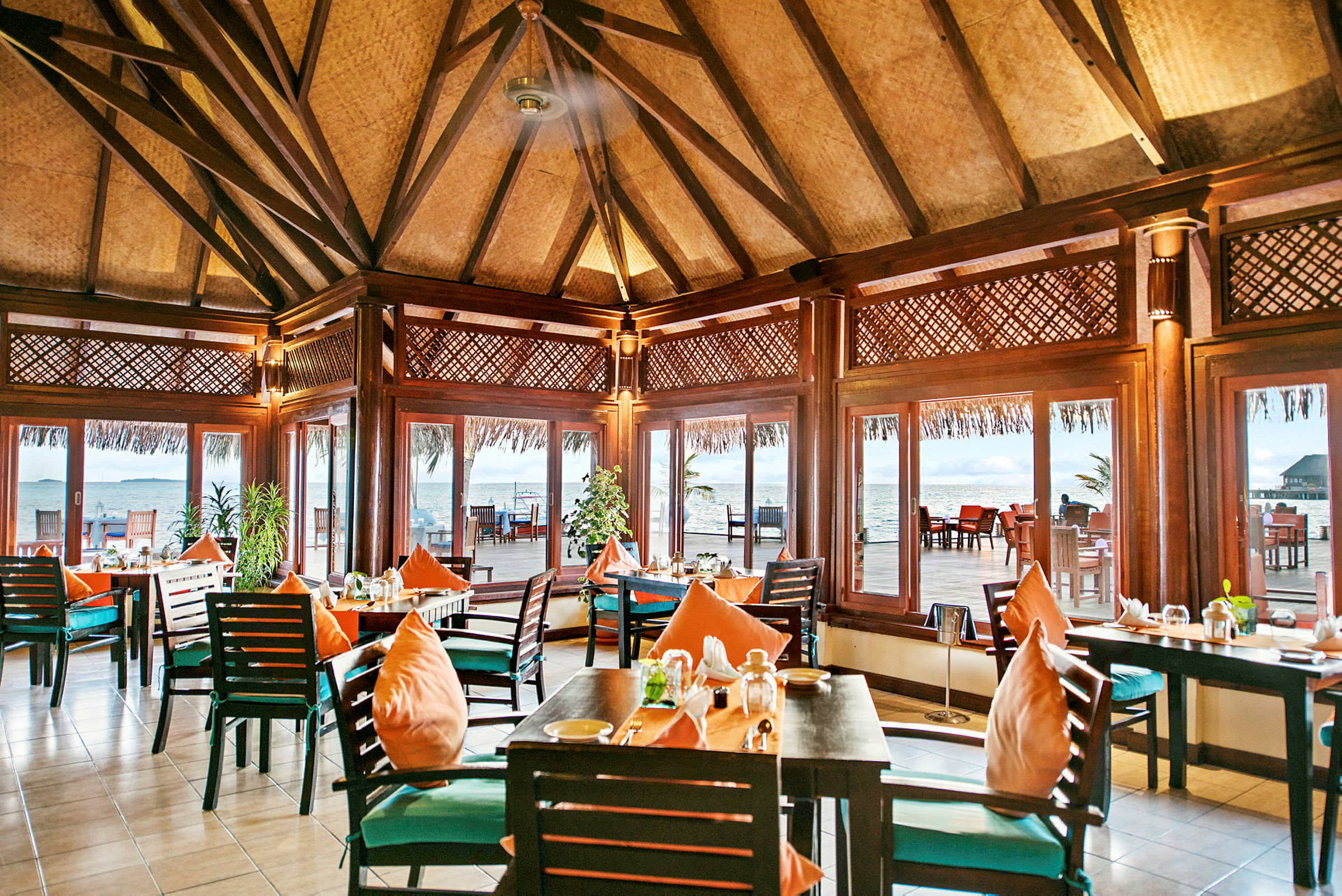 En restaurant med træinteriør og borde med orange puder, omgivet af store vinduer med udsigt over havet. (AI-genereret tekst)