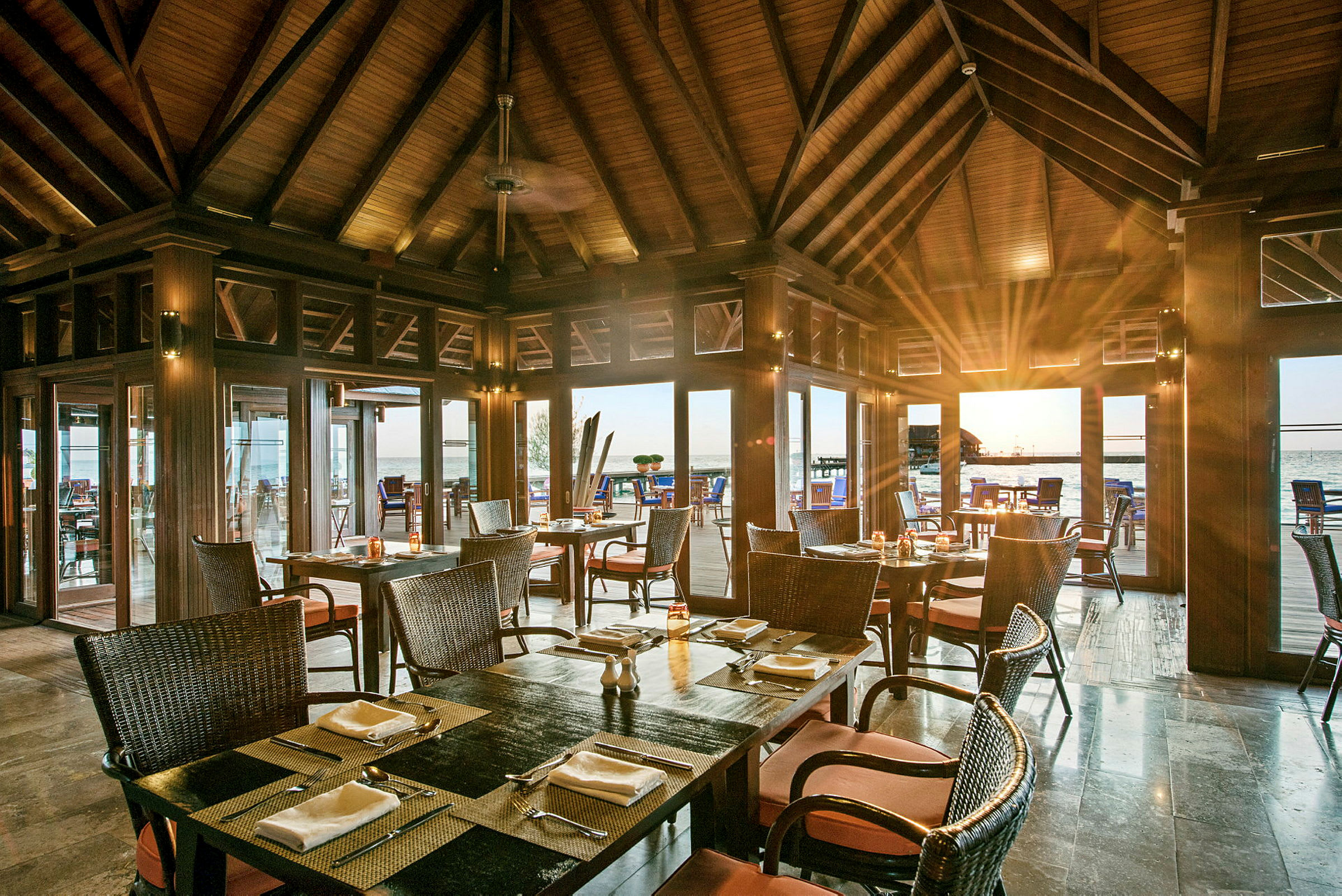 Restaurant med træindretning og dækkede borde, med udsigt til havet gennem glaspartier. (AI-genereret tekst)