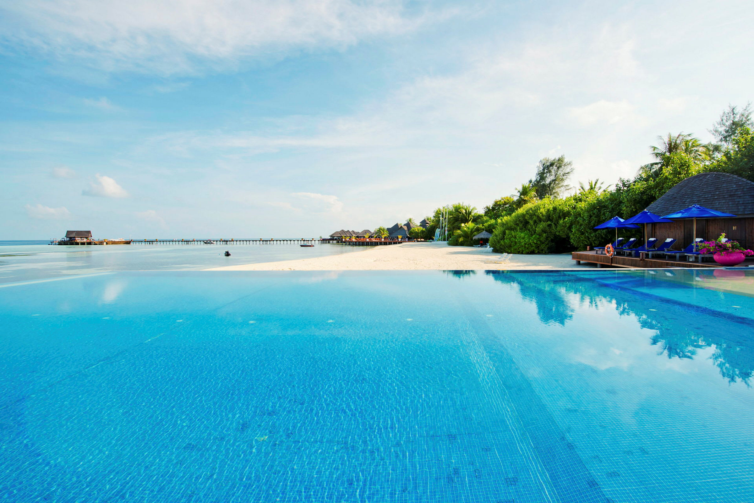 En swimmingpool med udsigt over stranden og havet, med liggestole og parasoller i baggrunden. (AI-genereret tekst)
