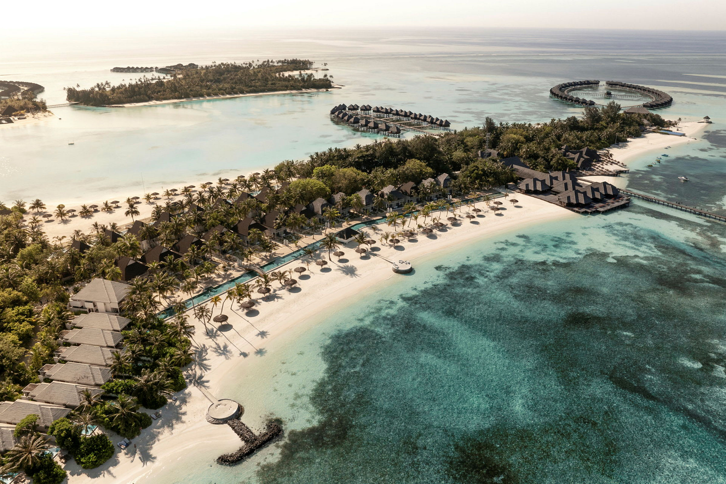 Luftfoto af en tropisk resort ved stranden med bungalows, swimmingpools og det omkringliggende klare hav. (AI-genereret tekst)