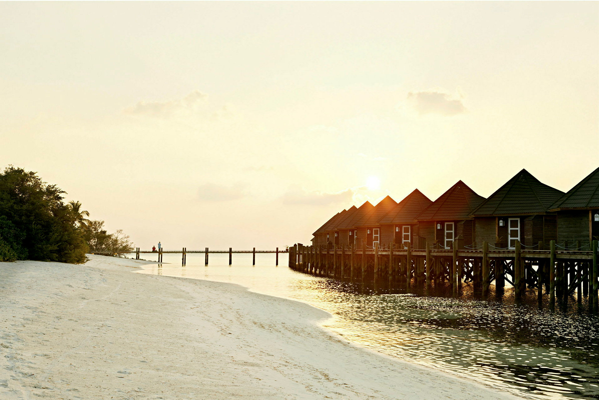 Bungalows på pæle over vandet ved en strand i solnedgang. (AI-genereret tekst)