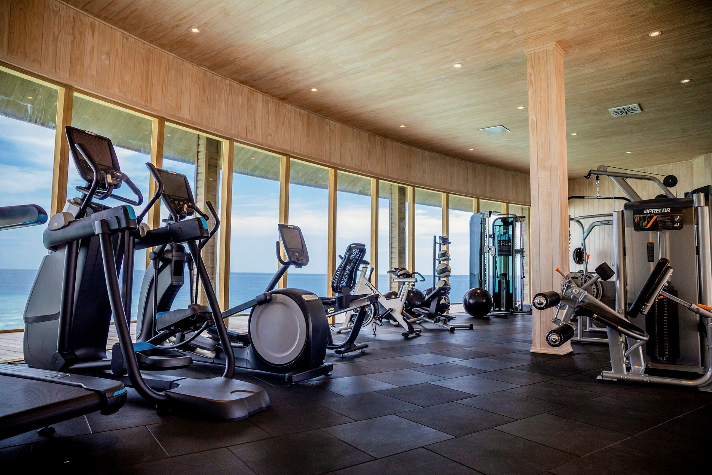 Et fitnesscenter med træningsmaskiner og store vinduer ud mod havet. (AI-genereret tekst)