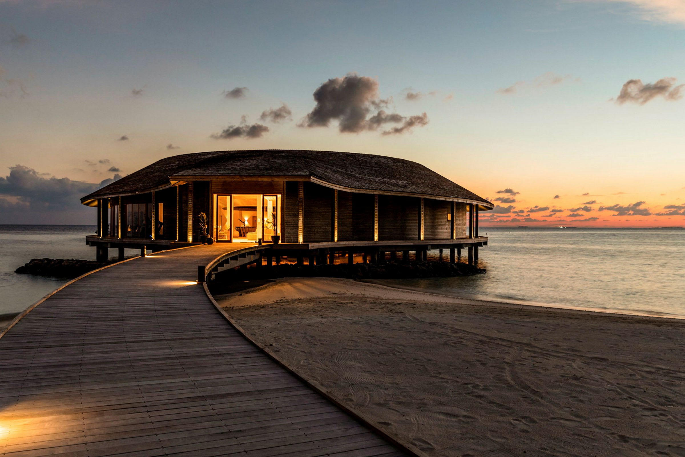 Et oplyst hus på pæle ved havet med en træsti, der fører frem til det, omgivet af sandstrand ved solnedgang. (AI-genereret tekst)